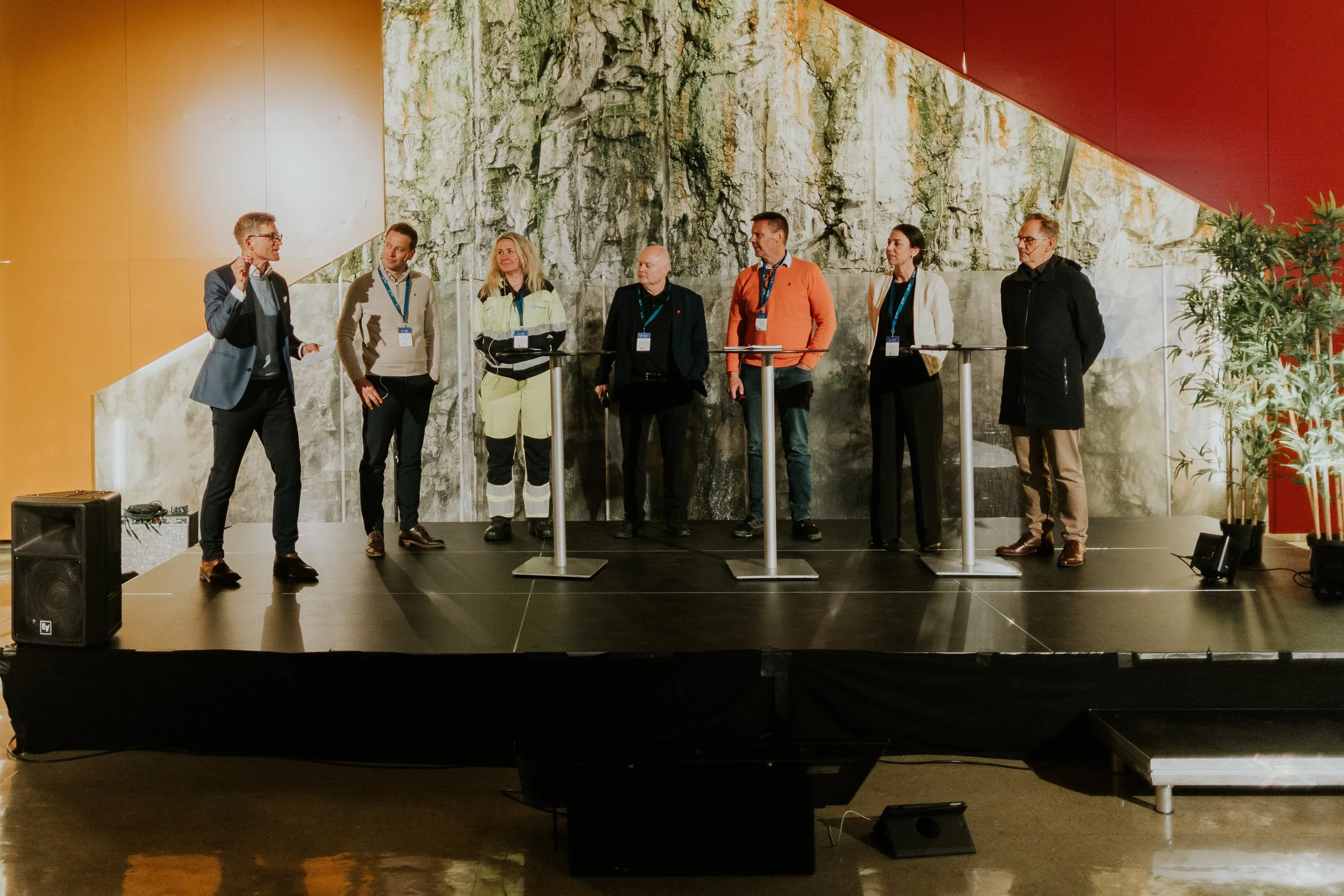 En gruppe på syv personer står på en liten scene med høytaler og mikrofoner, og diskuterer eller presenterer foran et abstrakt tre bakgrunn. Det er en voksen deltaker som snakker, mens de andre lytter oppmerksomt.