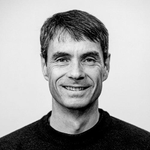 Et svart-hvitt bilde av en smilende mann med kort, brunt hår og en svart genser.