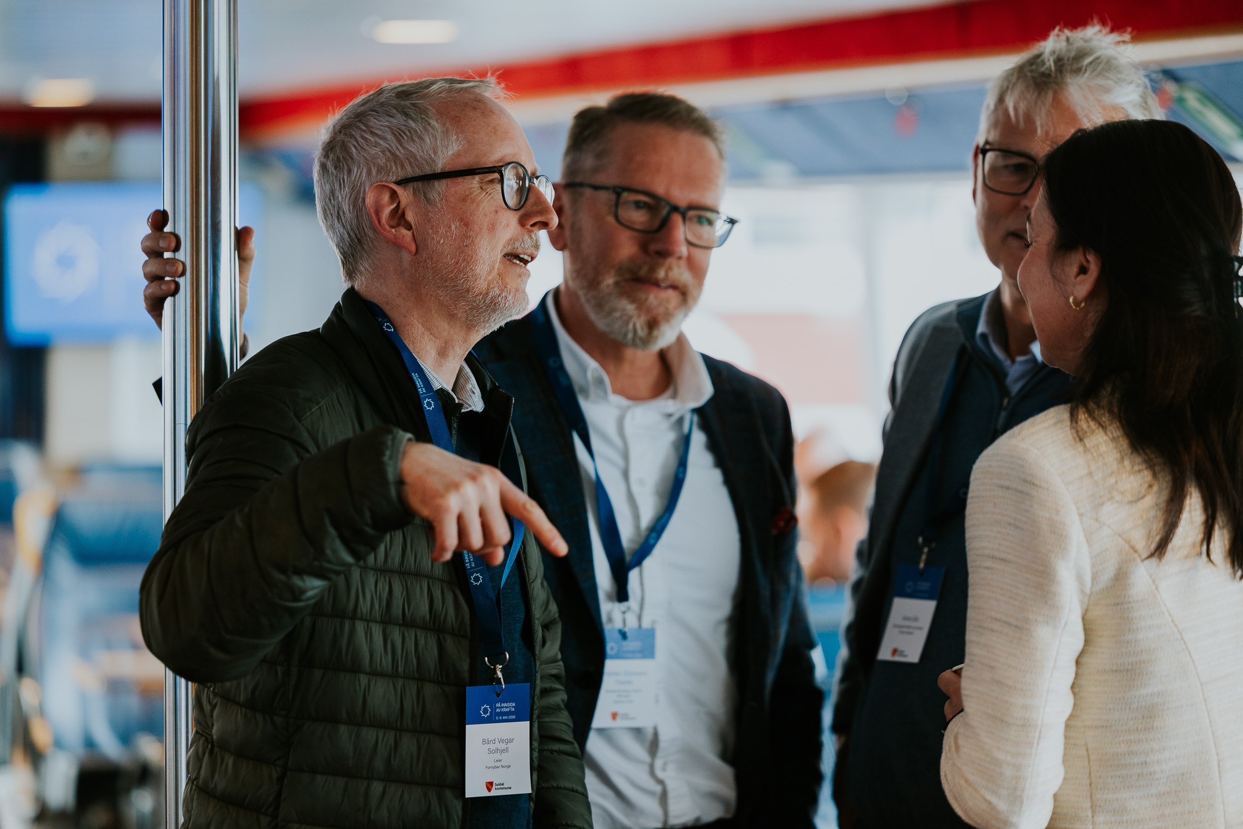 Fire personer i samtale på et messeområde, to menn med briller og en kvinne med mørkt hår. De har navneskiltene på halsen og er kledd i konferansetrekk.