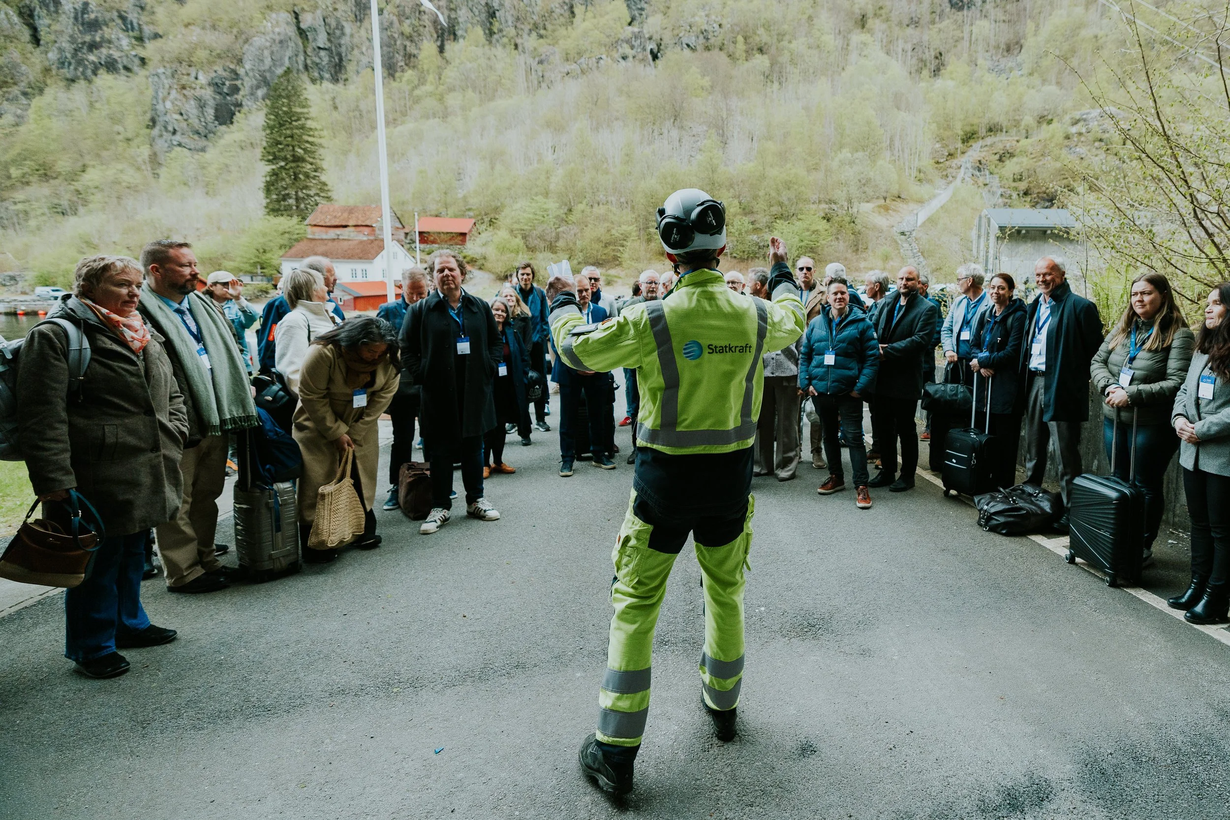 En gruppe mennesker som venter på en guide, mens en person i en høy synlig jakke med logoen Statkraft snakker til dem, i et naturskjønt område med fjell og hus i bakgrunnen.