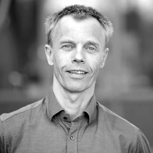 Portrett av en mann som smiler, kledd i en knappet skjorte, utendørs i svart-hvitt.