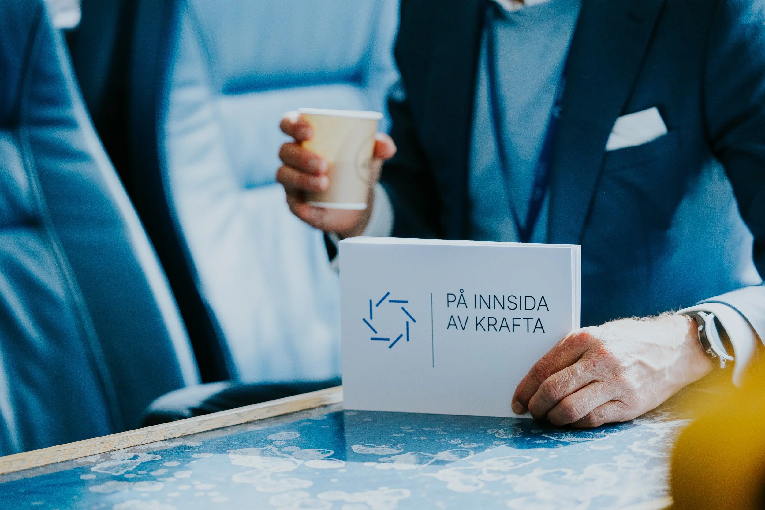 Nærbilde av en person som holder en beger med kaffe, omgitt av personer i formelle klær, på et arrangement.