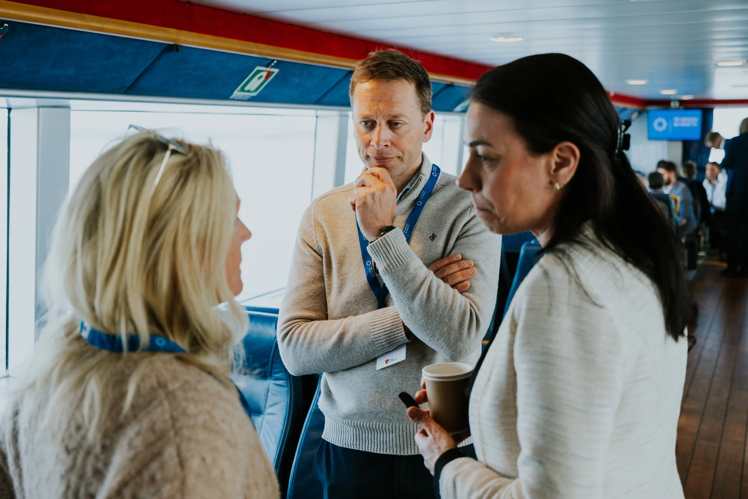 Tre personer som snakker i et terminalområde, med flyplassterminal i bakgrunnen, hvor flere mennesker venter.
