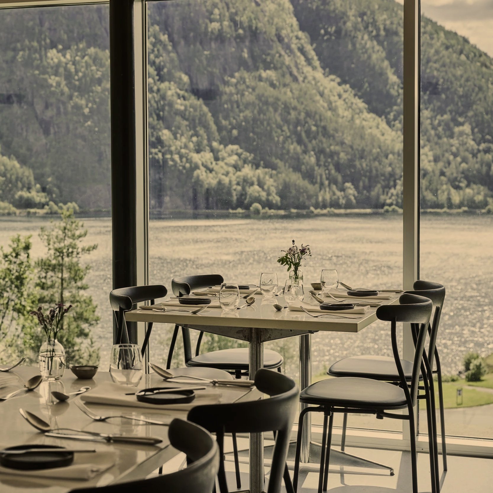 Restaurant med store vinduer som viser fjell og vann utenfor, med bord dekket med tallerkener, glass, bestikk og en liten blomsterbukett.