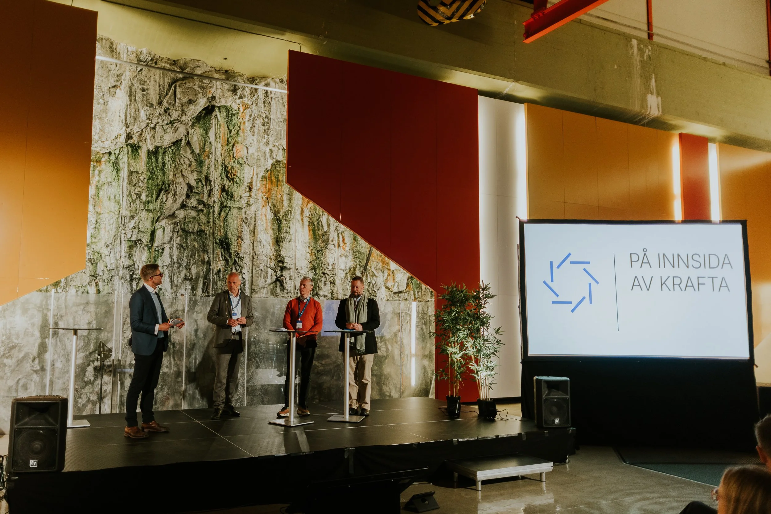Fire personer står og snakker på en scene. Bak dem er en vegg med en kunstnerisk stein- og marmorflate. Til høyre er et stort digitalt skjermbilde med teksten 'PÅ INNSIDA AV KRAFTA' og en grafisk logo.