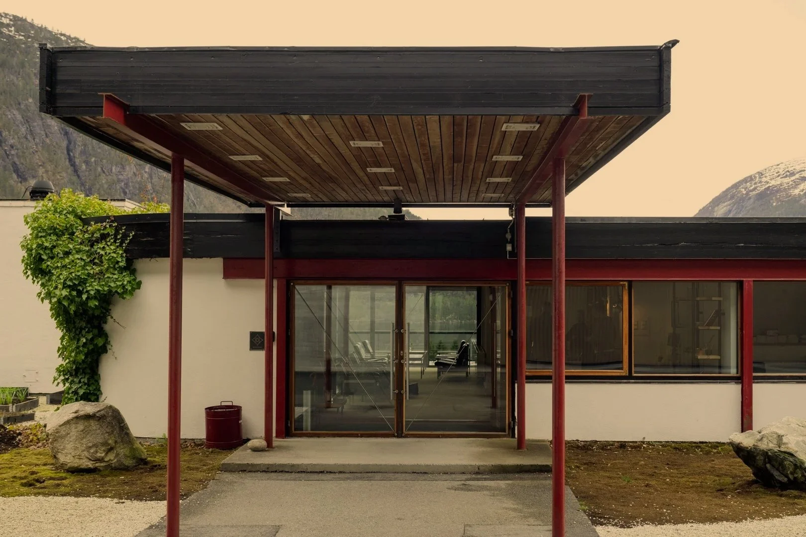 Modernt bygg med glassdører, redde og svarte vindusrammer, en utendørs overbygg støttet av røde stolper, og omgitt av fjell og landskap.