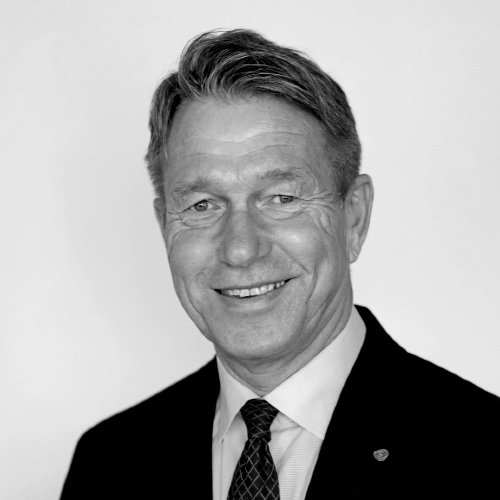 Portrett av en mann i svart dress og hvit skjorte, smilende, med kort, grått hår, i svart-hvitt.