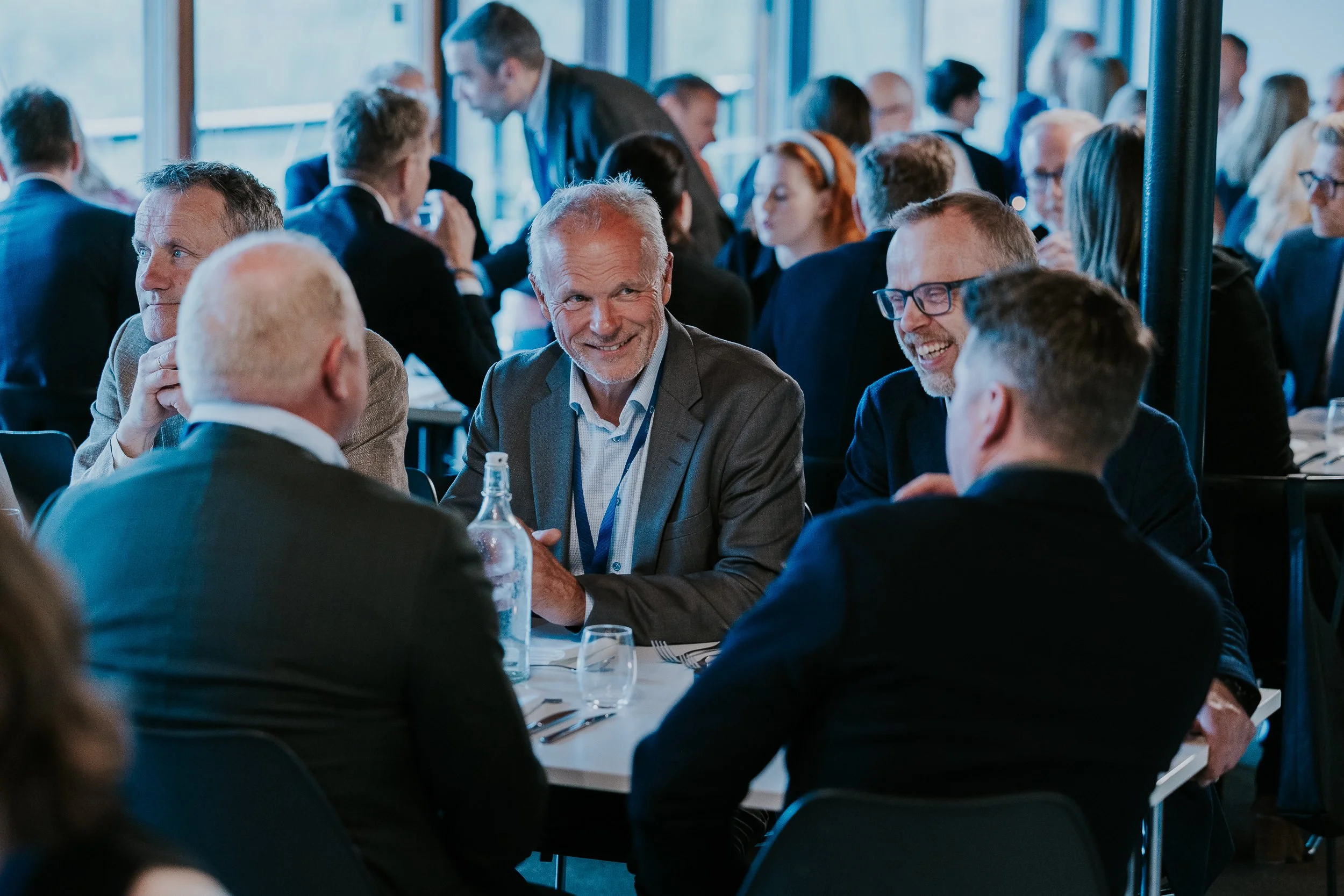 Mann med grå dress og hvitt skjegg smiler mens han pratar med andre personer ved et møtebord under en konferanse eller møte.