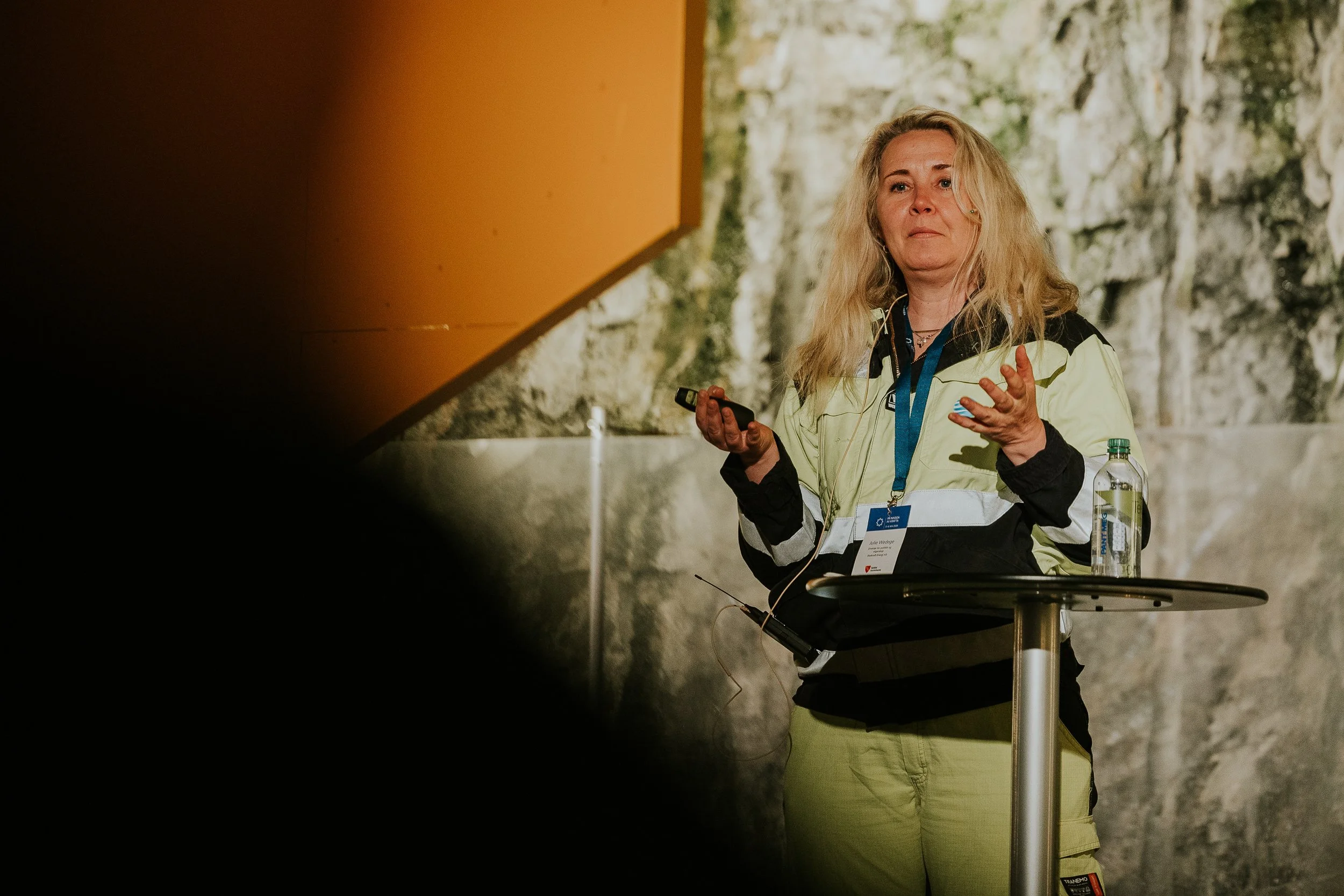 Kvinne foran en informasjonspult i en konsertsal, med rockbakgrunn, iført gul og svart jakke, og holder en mikrofon.