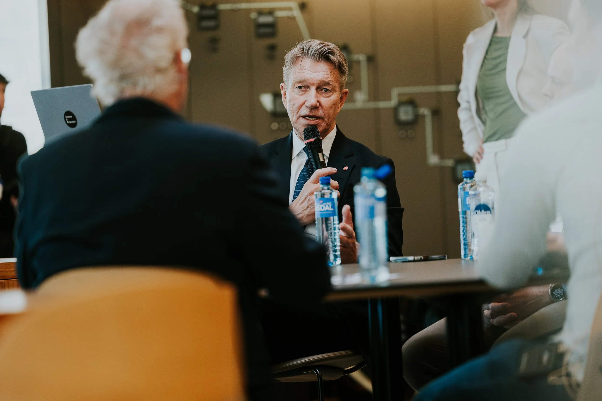 En mann med kort grått hår, iført mørk dress og hvit skjorte, snakker inn i en mikrofon under et møte eller paneldebatt. Han sitter ved et bord med vannflasker foran seg. Det er flere personer rundt ham, men de er delvis utenfor bildet, og bakgrunnen