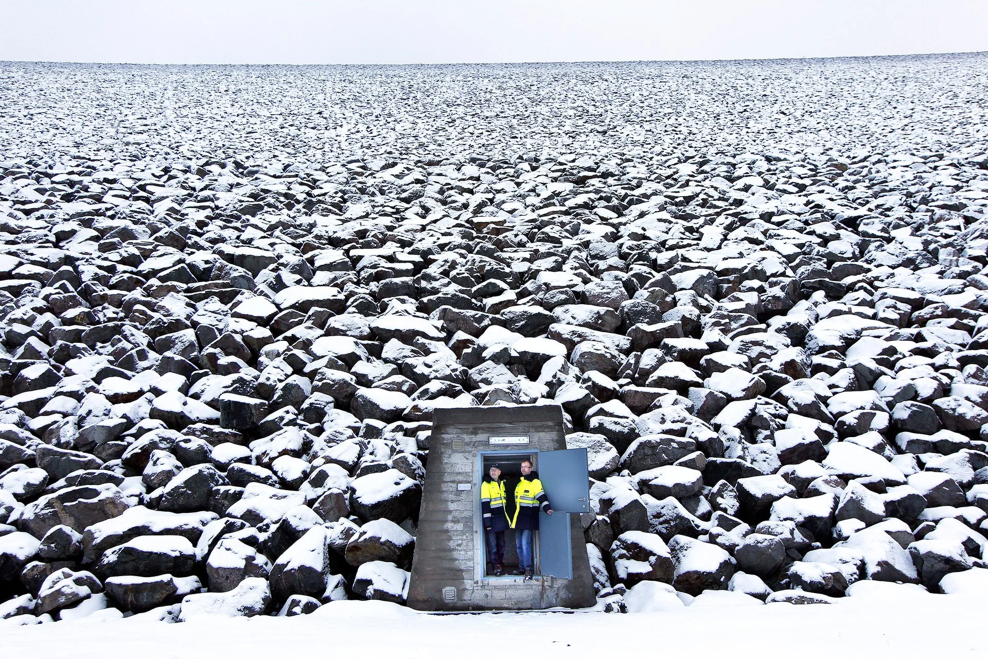 To personer i refleksvester står i en liten betongbygning med åpen dør, foran en steinbakke dekket av snø, med en isbre i bakgrunnen.