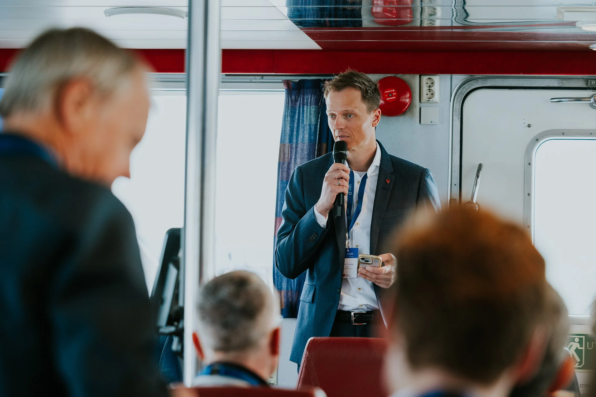 En mann i dress holder en mikrofon og snakker til passasjerer på et busstog.