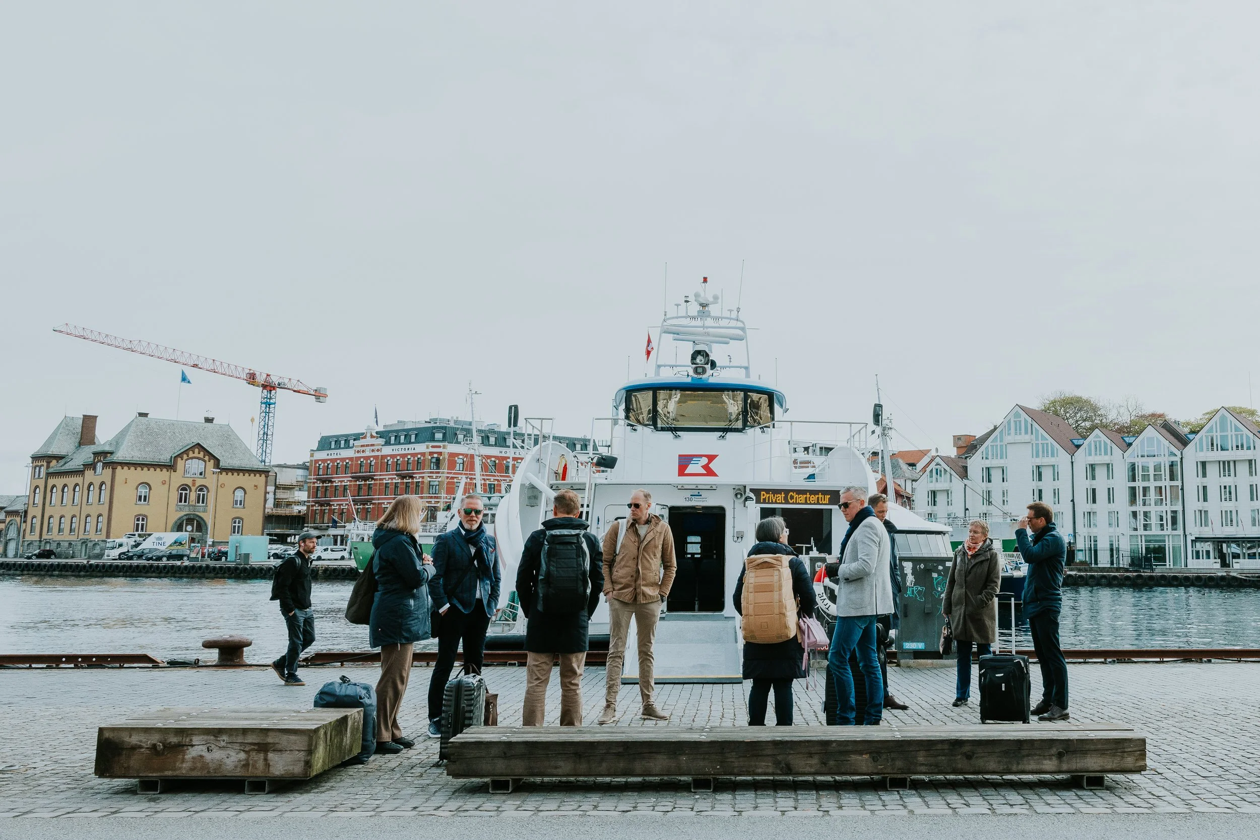 Grupper mennesker står og snakker foran en dykkerbåt ved havnen. Bygninger og en kran er i bakgrunnen.