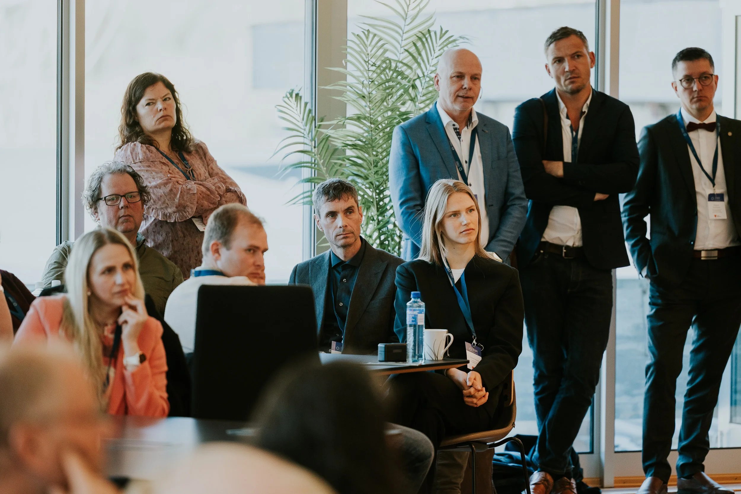 Grupp mennesker på et møte eller konferanse, noen står og noen sitter, innendørs med store vinduer og plante.