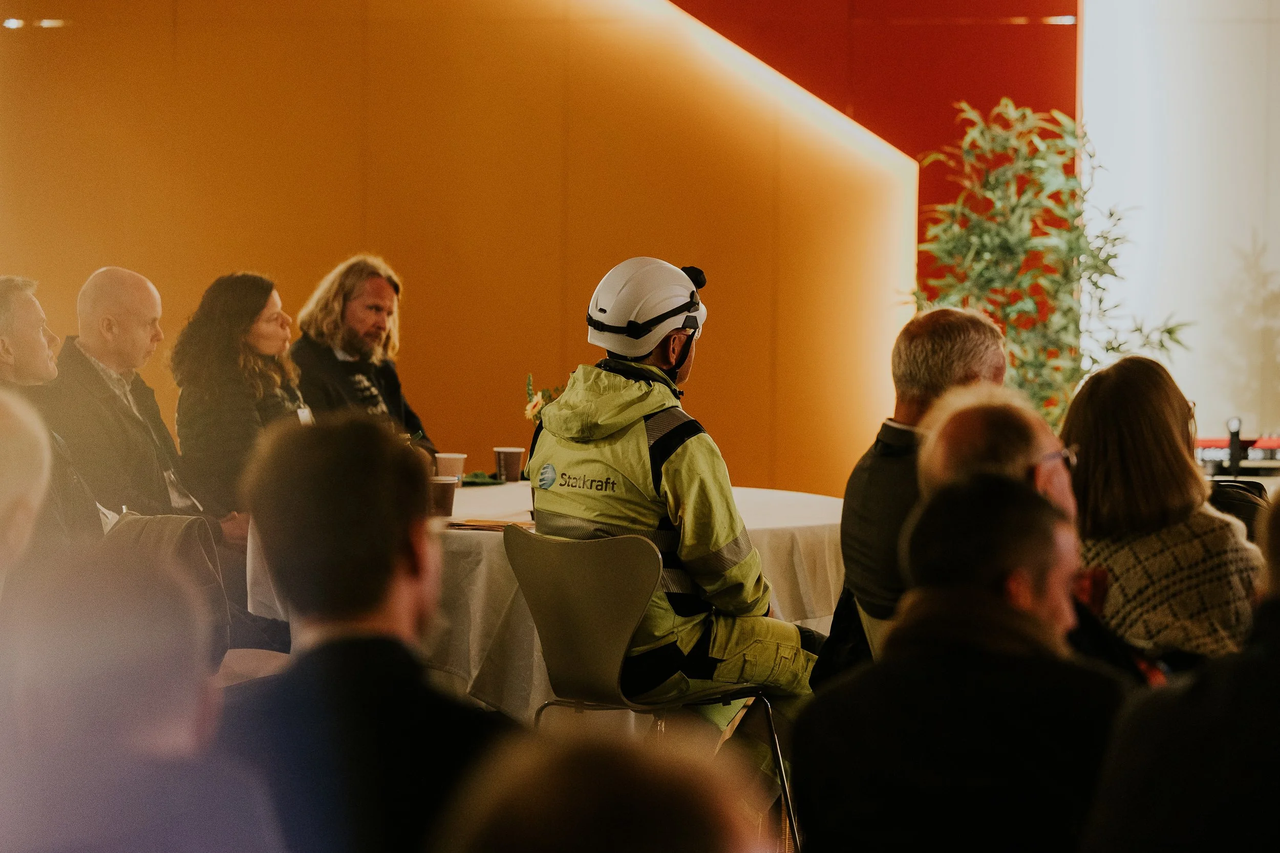 Mennesker sitter i et auditorium med oransje og hvit vegg, blant dem en person i gult arbeidsantrekk med hjelm med logo 'Statkraft'.