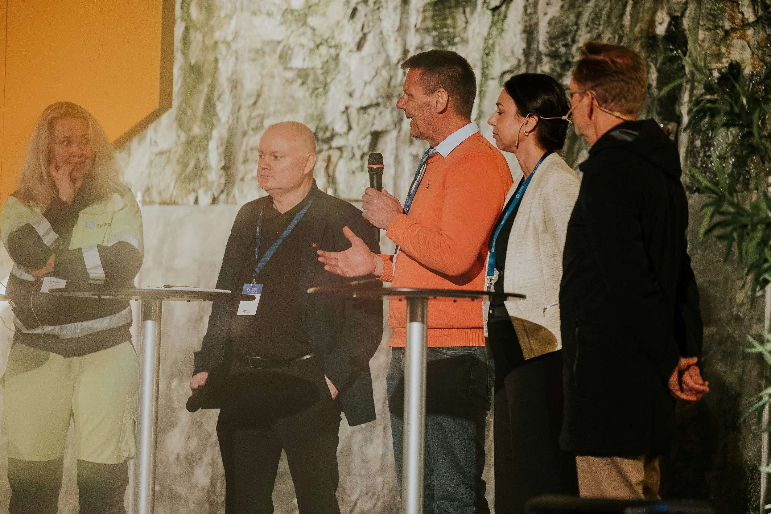 En gruppe mennesker deltar i en diskusjon eller panel, hvor en mann holder en mikrofon, mens de andre lytter oppmerksomt. De står foran en steinmur eller naturbakgrunn.