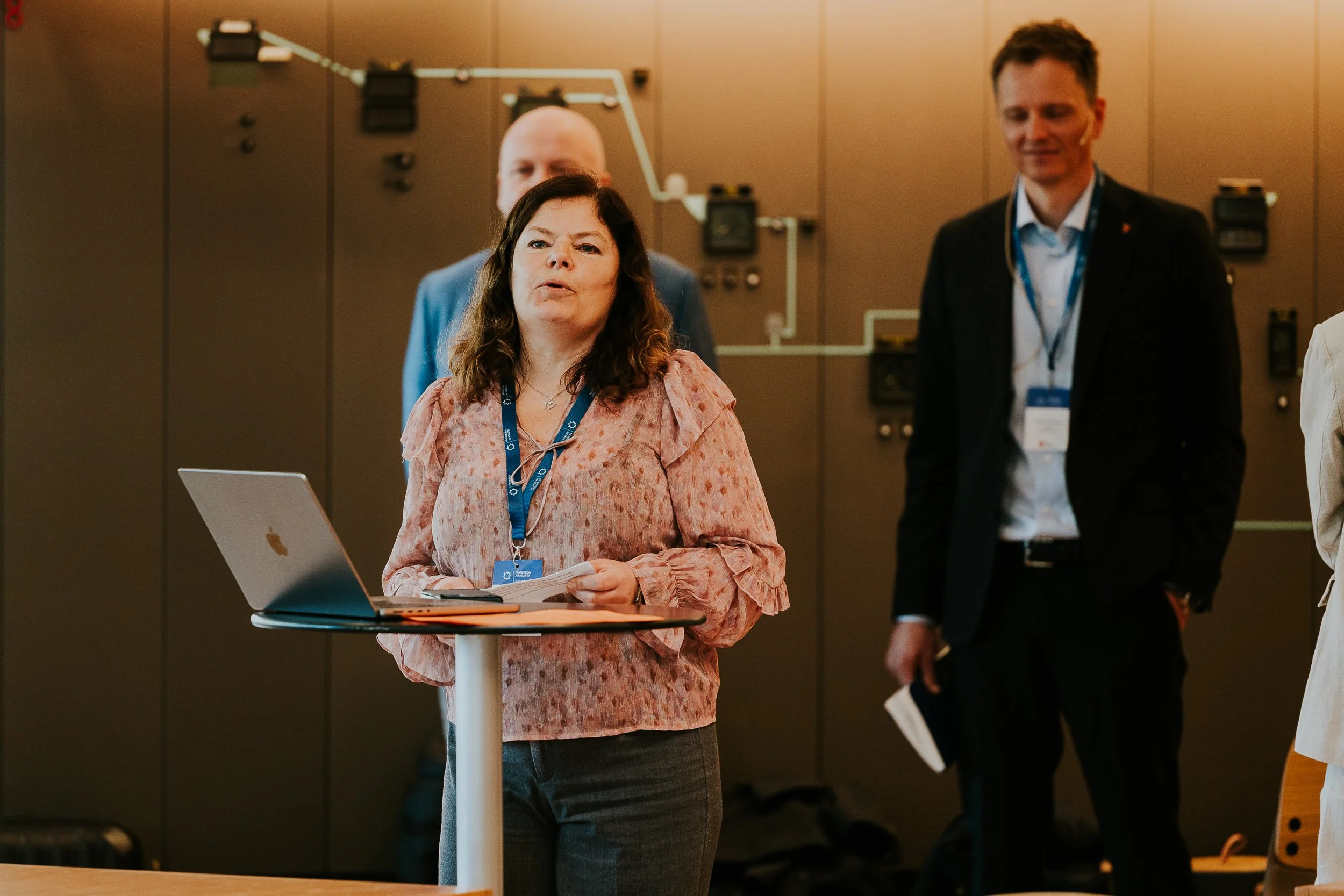 Kvinne med brunt hår og rosa bluse står foran en laptop og snakker, mens menn i bakgrunnen ser på og lytter på et innendørs konferanseseminar.
