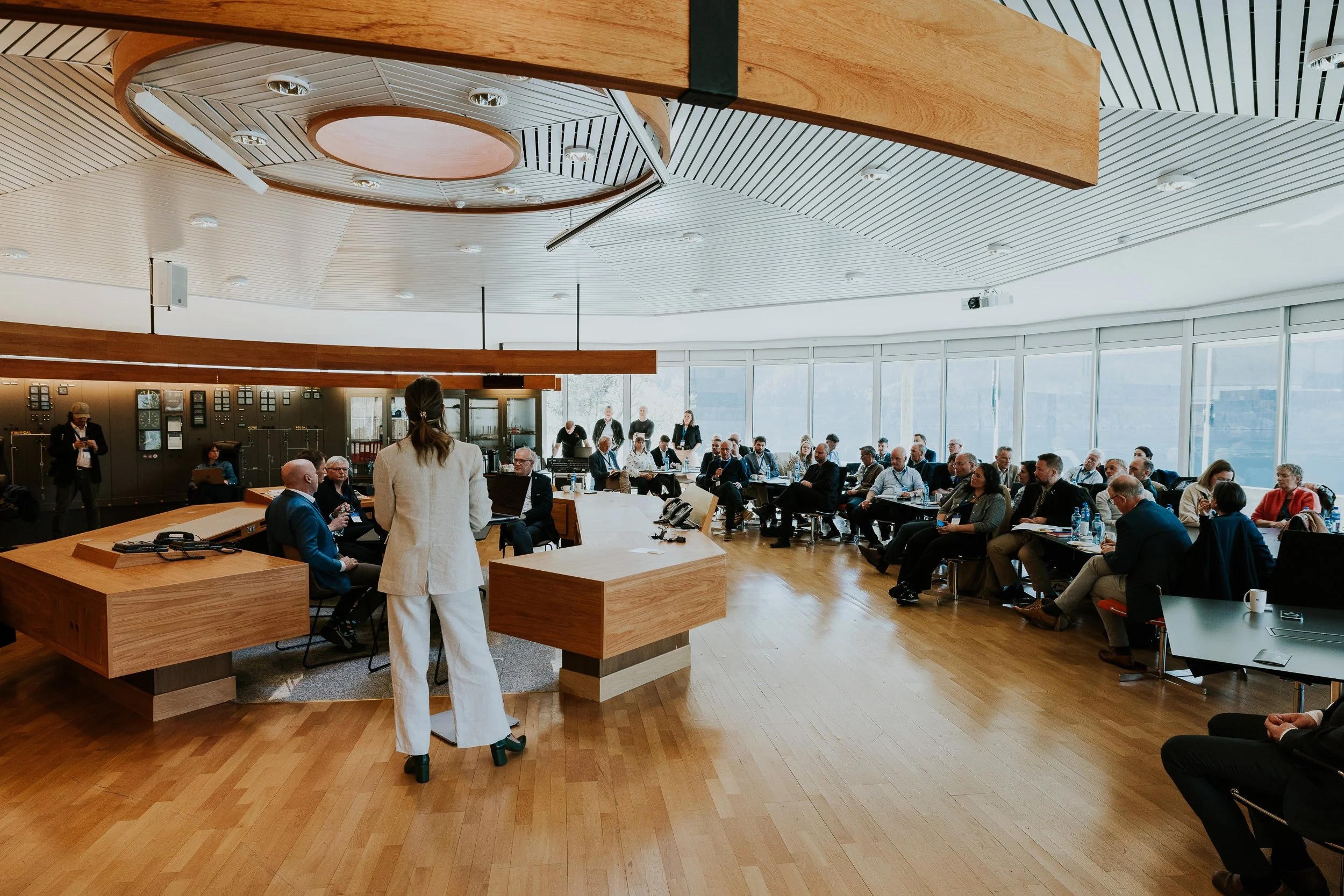 Et møte eller konferanse med mange deltakere sittende i en stor, moderne konferansesal med store vinduer og panoramautsikt. En person står foran gruppen, sannsynligvis som presenterer eller snakker.