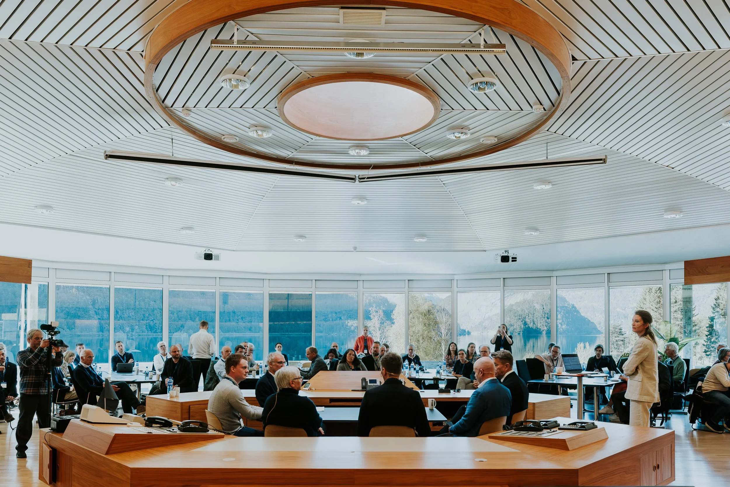 Et møte i et konferanserom med flere personer som sitter rundt langbord, mens en kvinne står ved enden av rommet og snakker. Rommet har store vinduer og et designet tak med lys og trepanel.