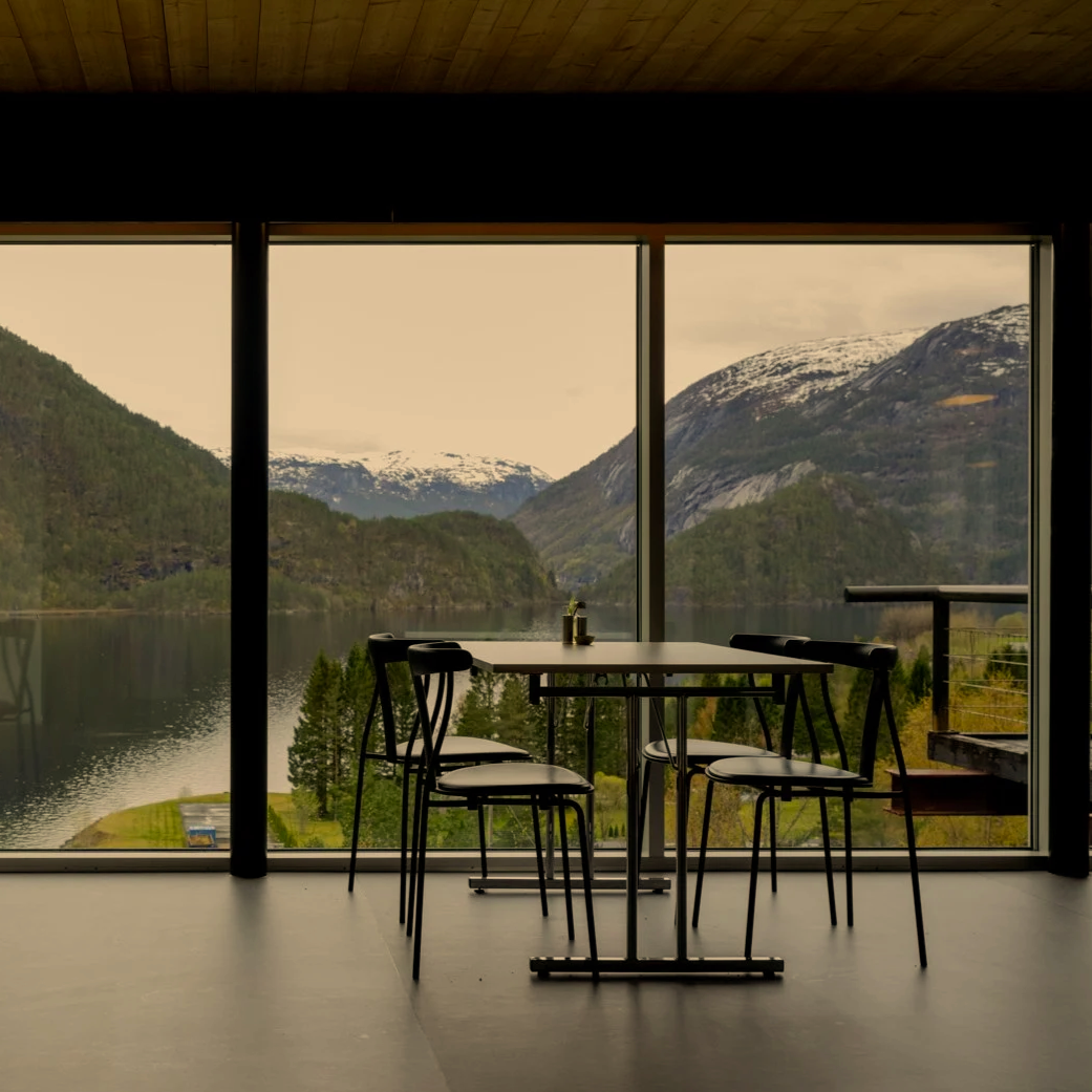 Innendørs bilde av et lite spisebord med fire stoler foran store vinduer som viser fjell, elv og skog i nabolaget.