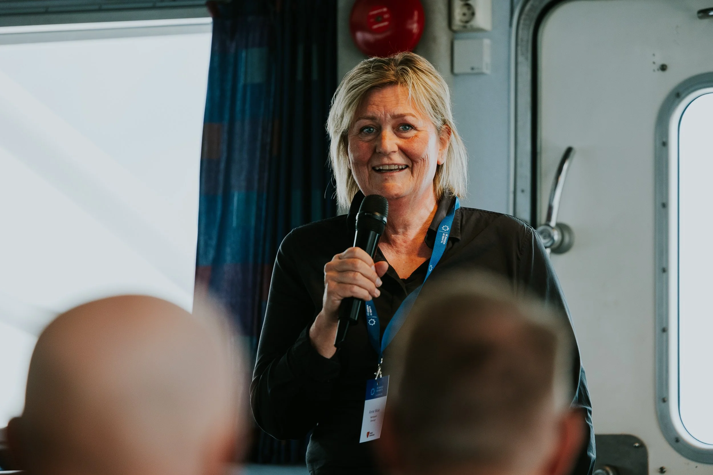 En kvinne med blond hår holder en mikrofon og snakker, mens hun står i et rom med en dør og en brannalarm på veggen.