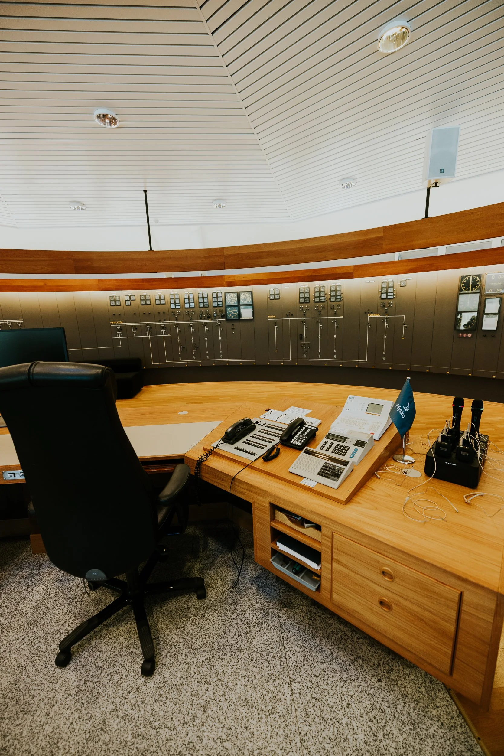 Kontor med et rundt trebord med telefoner, datamaskiner, dokumenter og en liten flagg, med svarte kontorstoler og en stor trekommode med skuffer. Bak står en vegg av kontrollpaneler og utstyr.