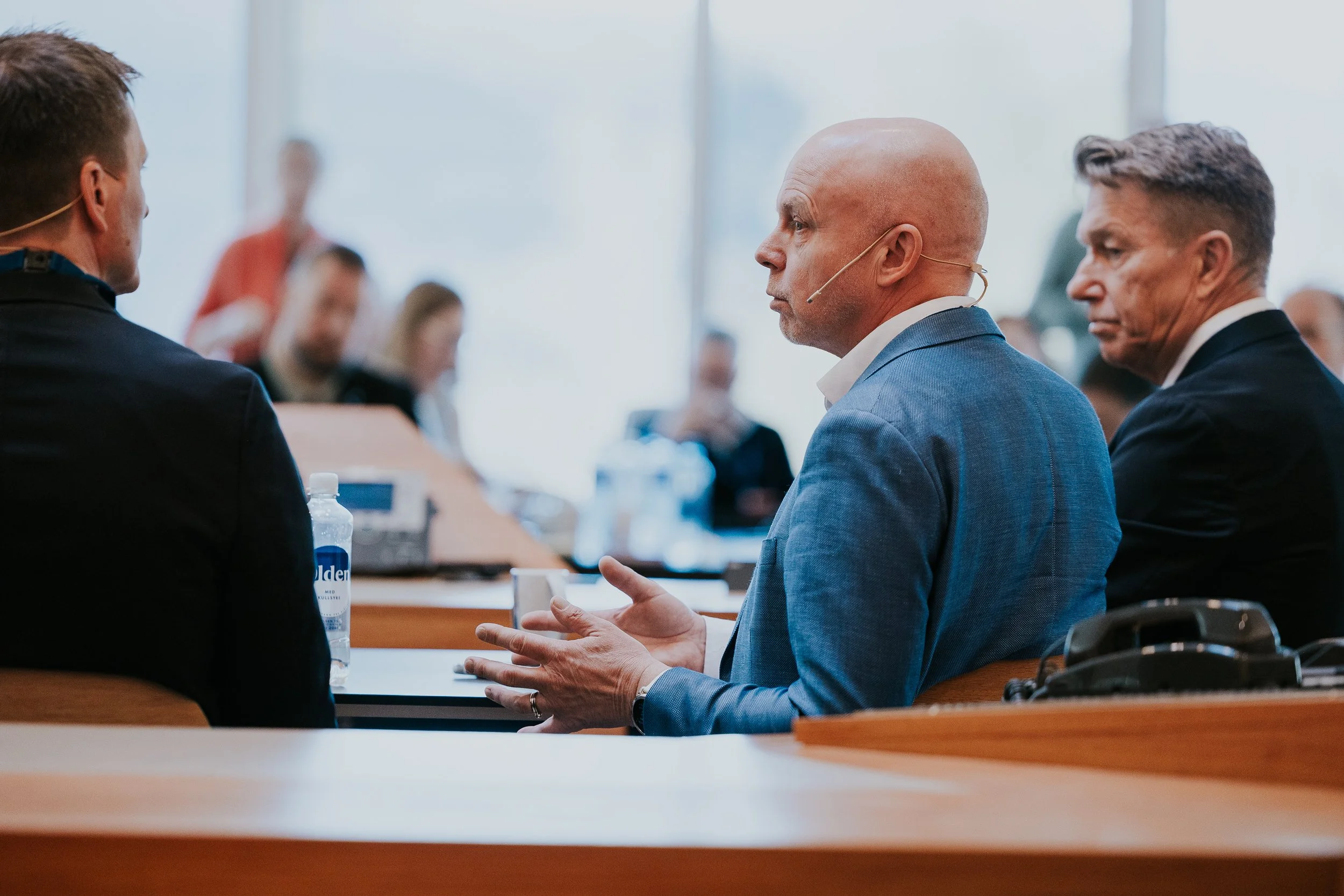 Tre personer i et møte, to er tydelig sett i fokus, en med blå dress og en med svart dress. De snakker og har notater foran seg.