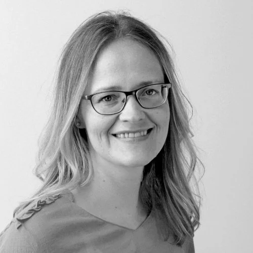 Et svart-hvitt portrett av en smilende kvinne med briller og skulderlangt hår.