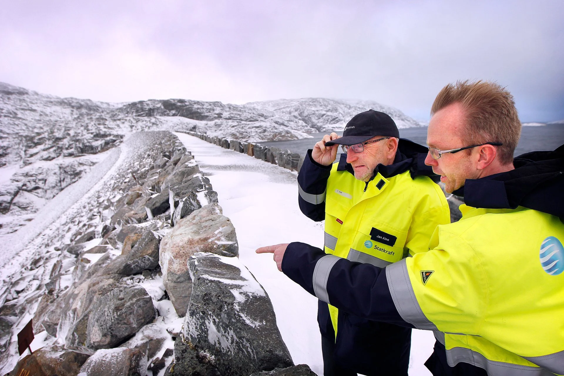 To menn i gult vintertøy ser på en steinmur de er ved å undersøke i en snødekt, fjellrik landscape.