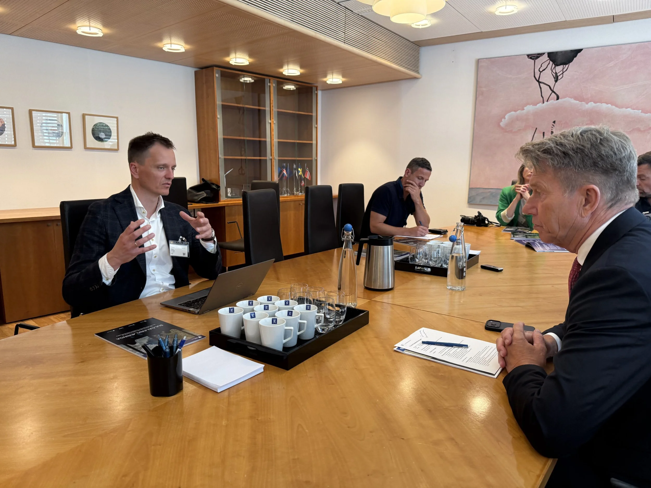 Fem personer deltar i et forretningsmøte i et konferanserom. En mann i dress snakker gestikulerende, mens de andre lytter og tar notater. Bordet har kopper, vann og dokumenter.