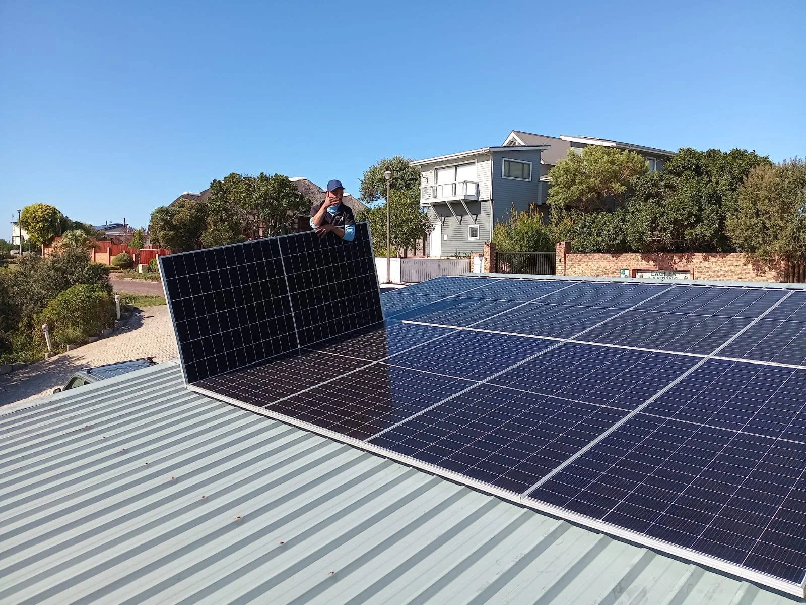 A man standing on a rooftop with solar panels, touching his face, in a residential neighborhood with houses and trees under a clear blue sky.