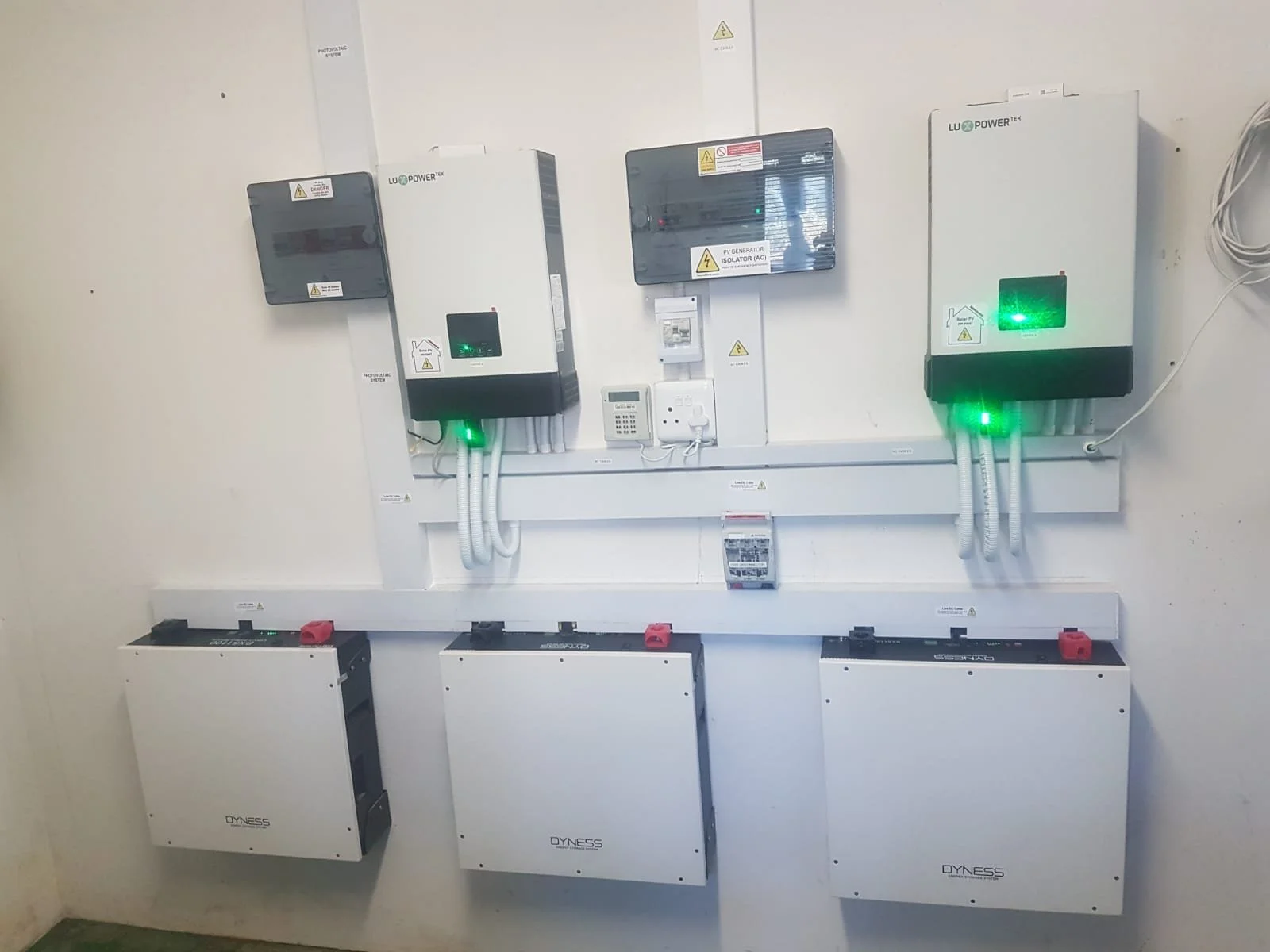 Electrical equipment and solar inverters mounted on a white wall, with green indicator lights, cables, and labels.