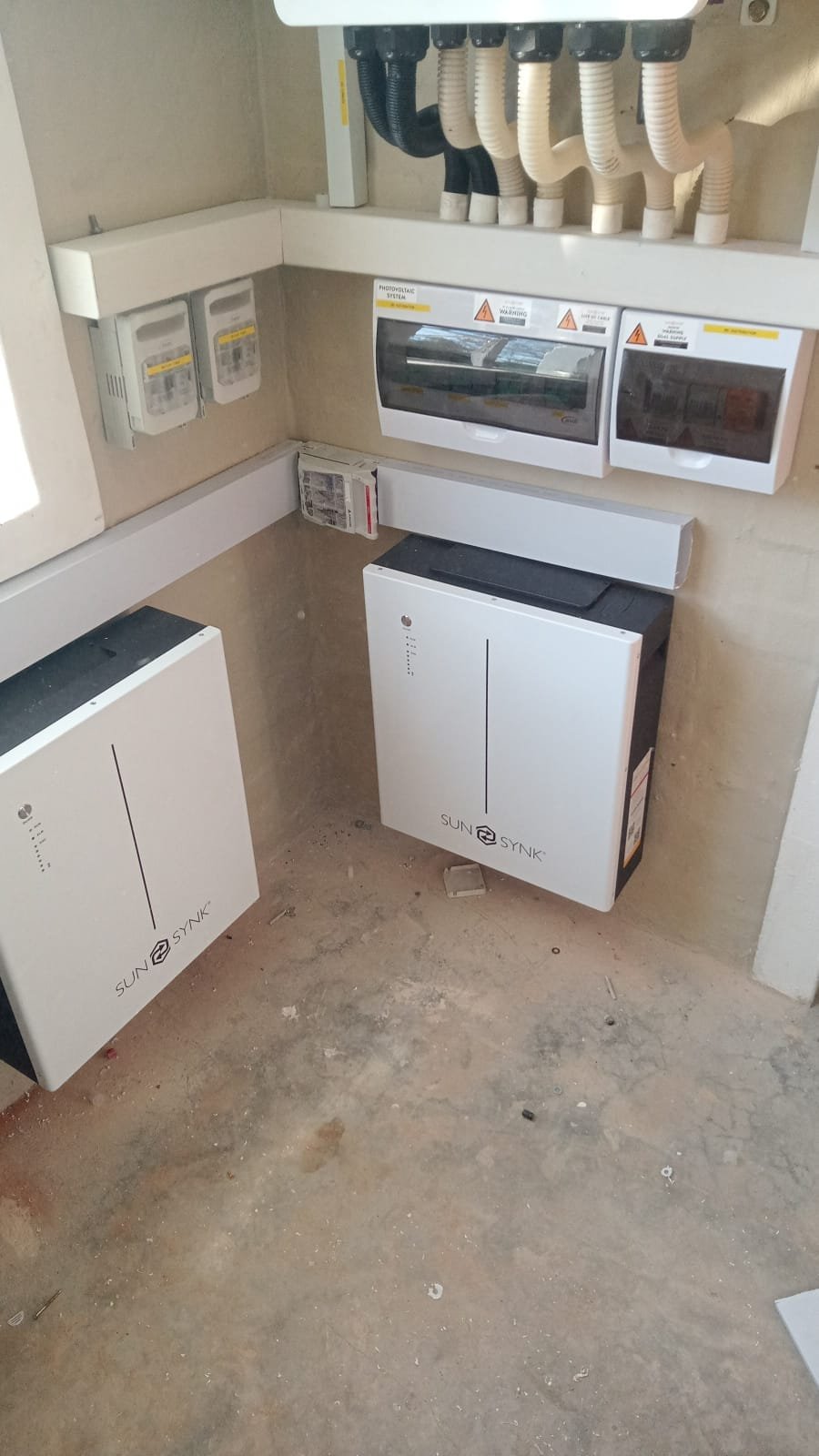 Electrical and utility boxes, circuit breakers, and conduit pipes mounted on a beige wall in a utility or breaker room.