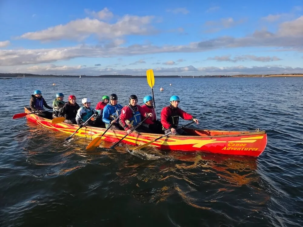 A group of ten people in a colorful canoe on a body of water, wearing helmets and life jackets, with a clear sky and distant shoreline in the background.
