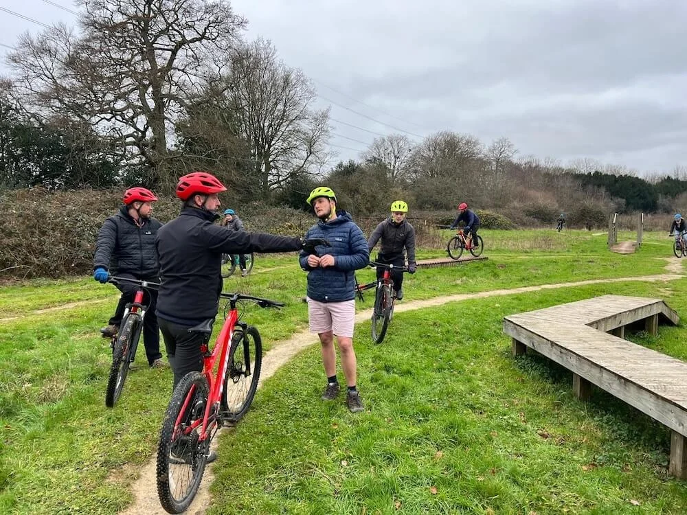 Group of people wearing helmets and jackets with bikes, standing on a grassy trail in a park, with some riding bikes in the background.