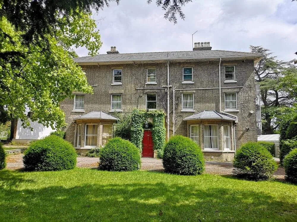 Burwell House - A large, three-story brick house with a red door, surrounded by green bushes and trees, under a partly cloudy sky.