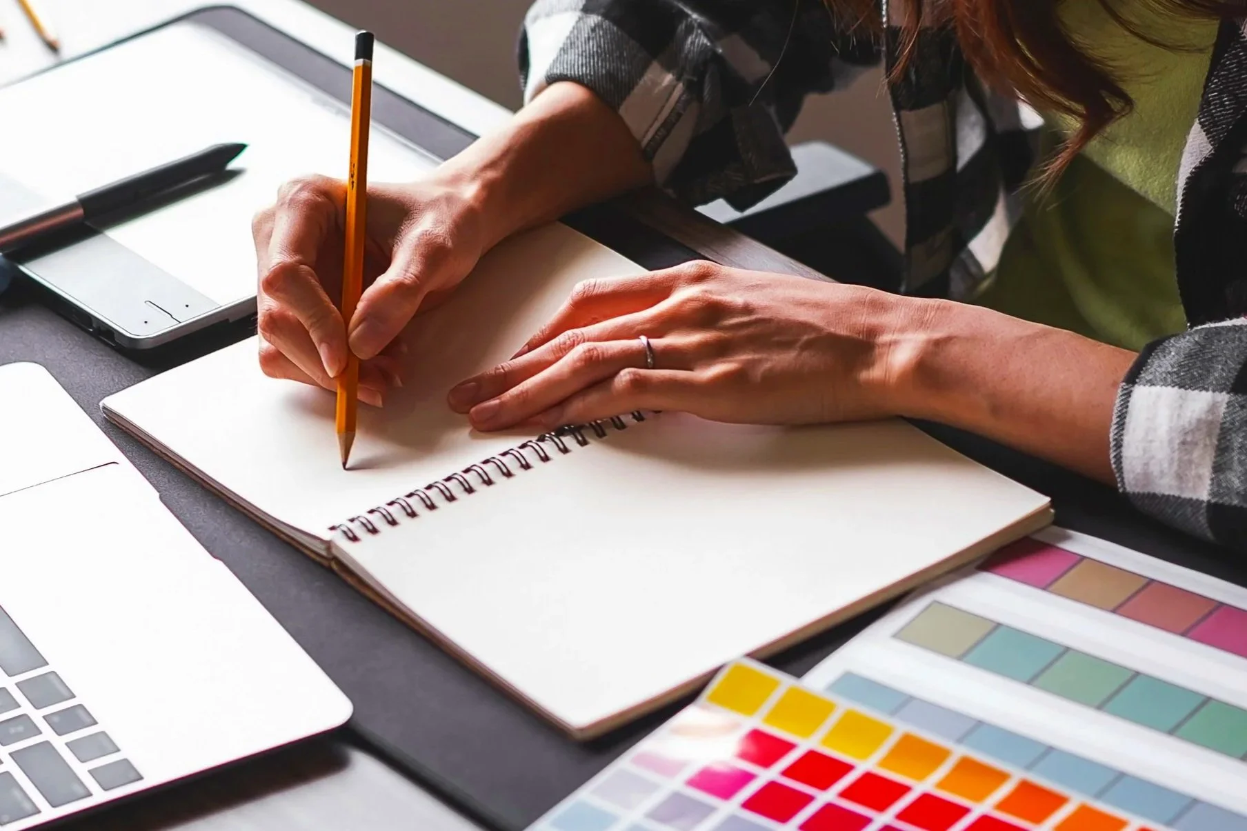 Une personne écrit dans un carnet à spirale sur un bureau avec un stylo, un ordinateur portable, des échantillons de couleurs et une tablette graphique autour.