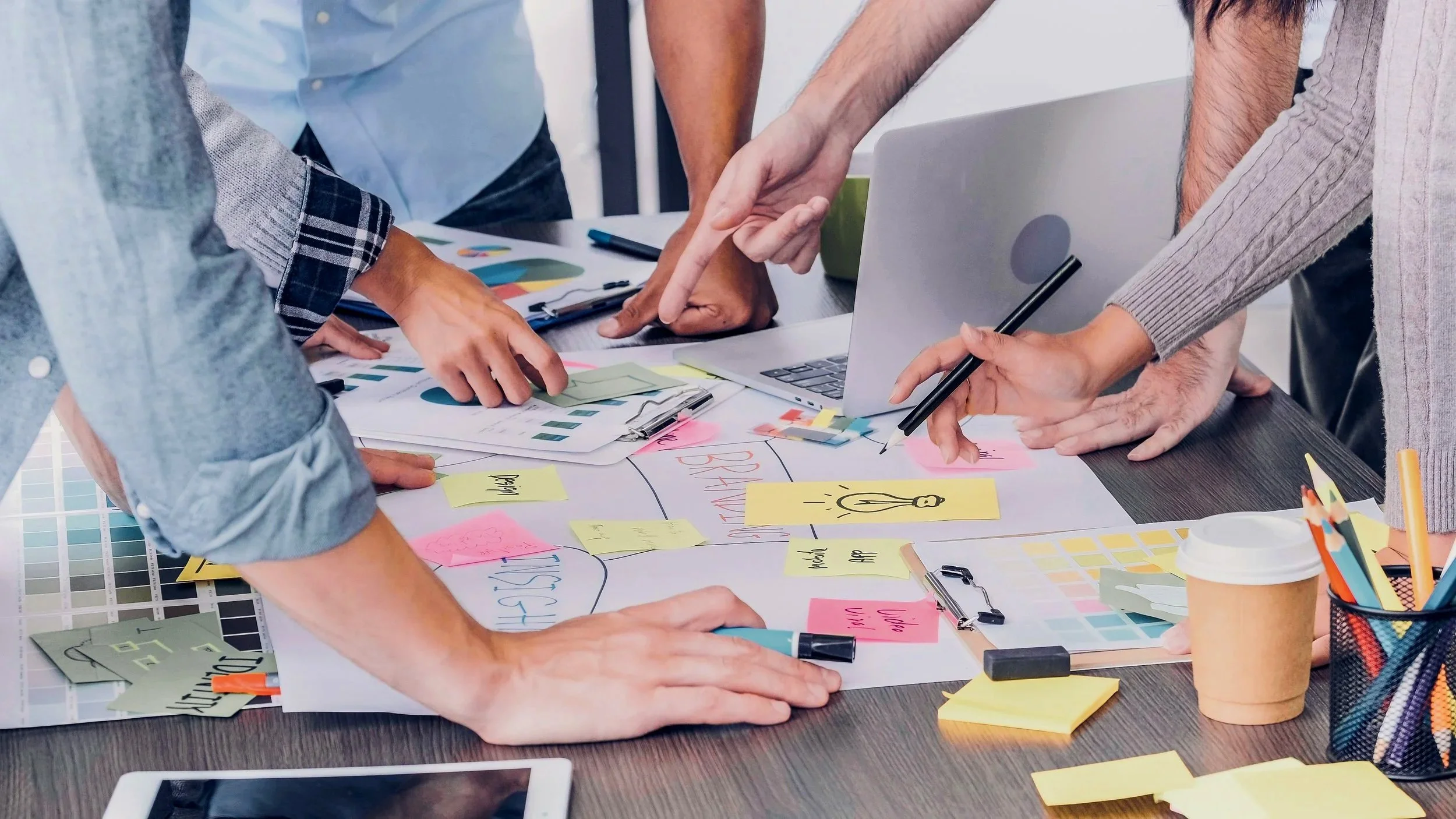 Groupe de personnes en réunion autour d'une table, discutant de plans et d'idées, avec des notes adhésives, un ordinateur portable, des stylos, et un café.