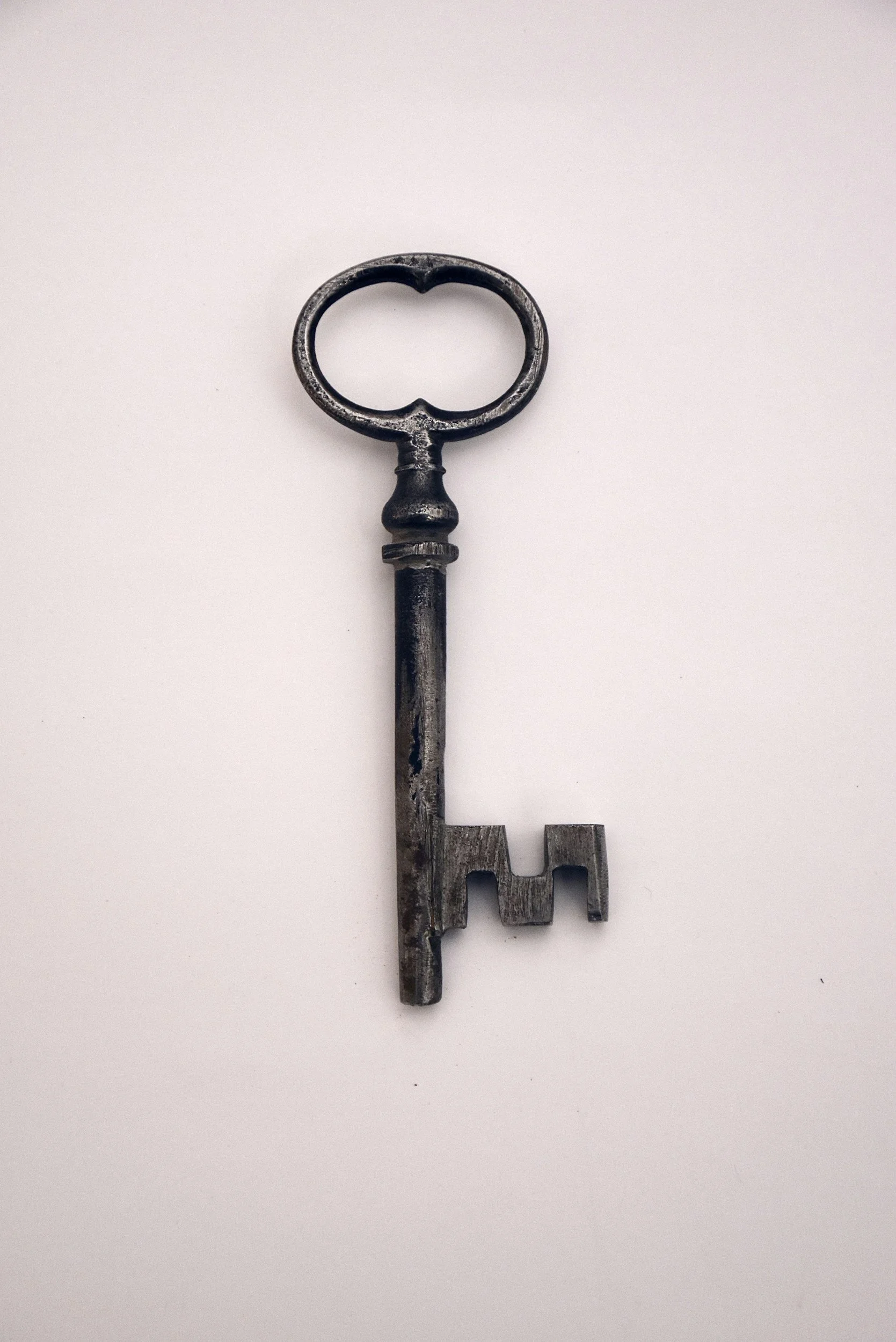 An antique metal key with a round handle and a complex bit, lying on a plain white surface.