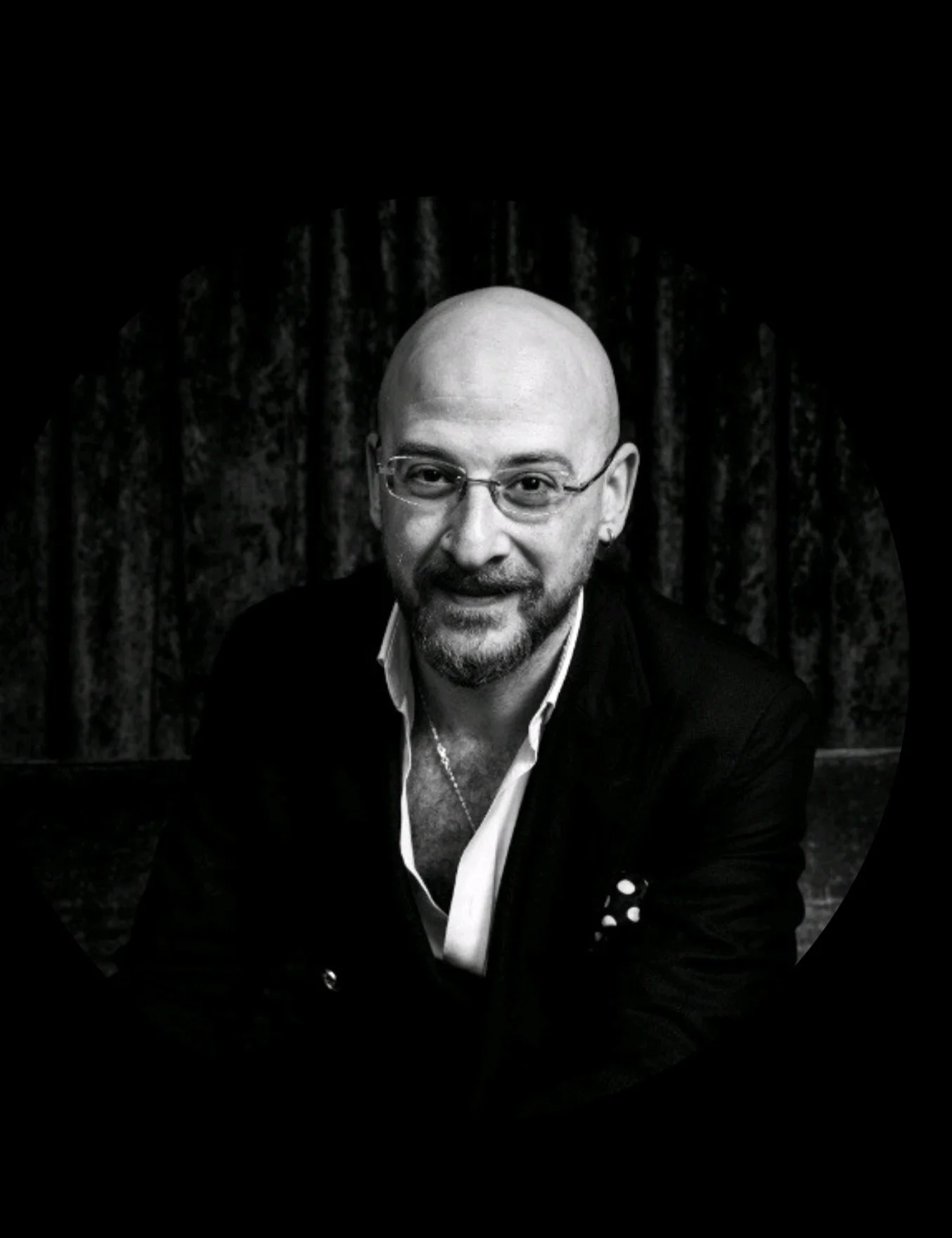 Black and white portrait of a bald man wearing glasses, a white shirt, and a dark blazer, smiling at the camera in front of a curtain background.