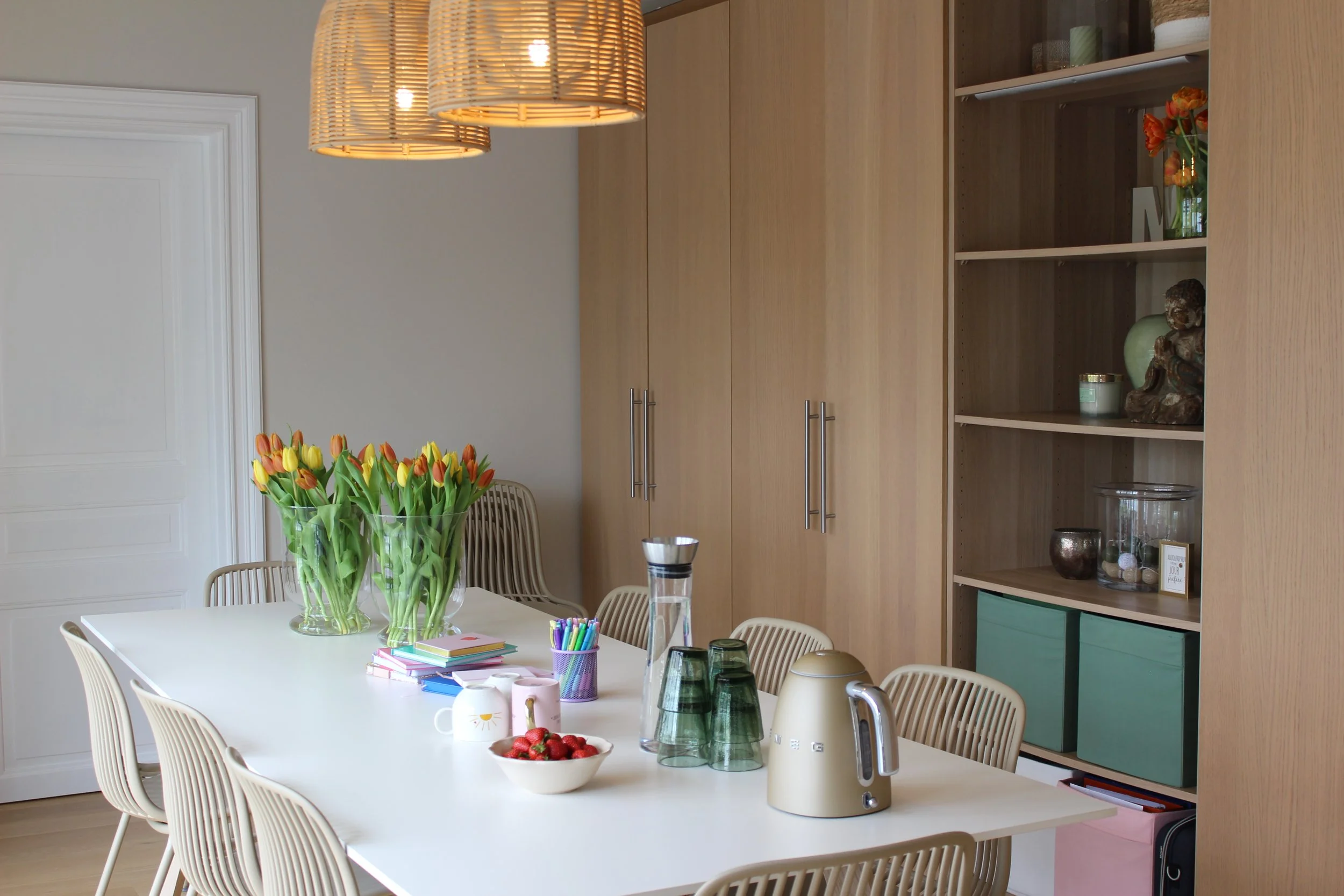 Salle de réunion avec une table blanche, un vase avec des tulipes, plusieurs tasses, un bol de fraises, une bouteille d'eau et une armoire en bois clair avec des objets décoratifs.