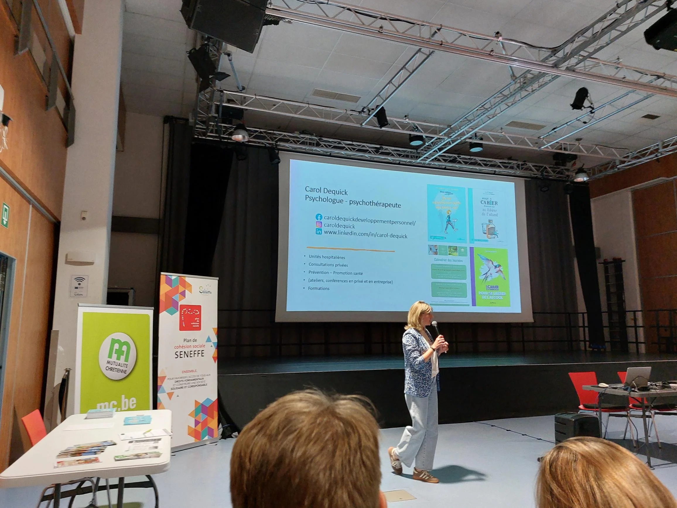 Une femme parle avec un microphone sur scène, projection affichée en arrière-plan avec ses coordonnées, dans une salle de conférence ou d'événement.