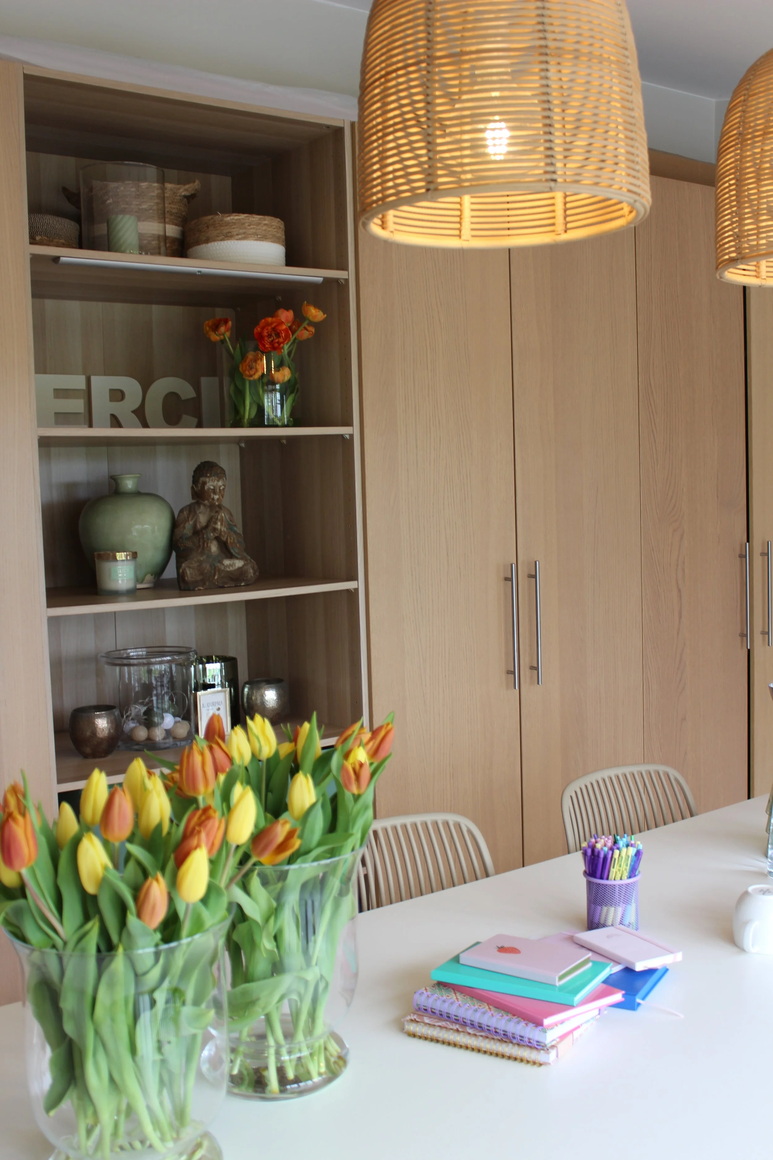 Un vase avec des tulipes jaunes et orange sur une table blanche, plusieurs carnets colorés et stylos violets dans un porte-stylos violet, et un mur avec des étagères en bois avec des décorations et des ornements.