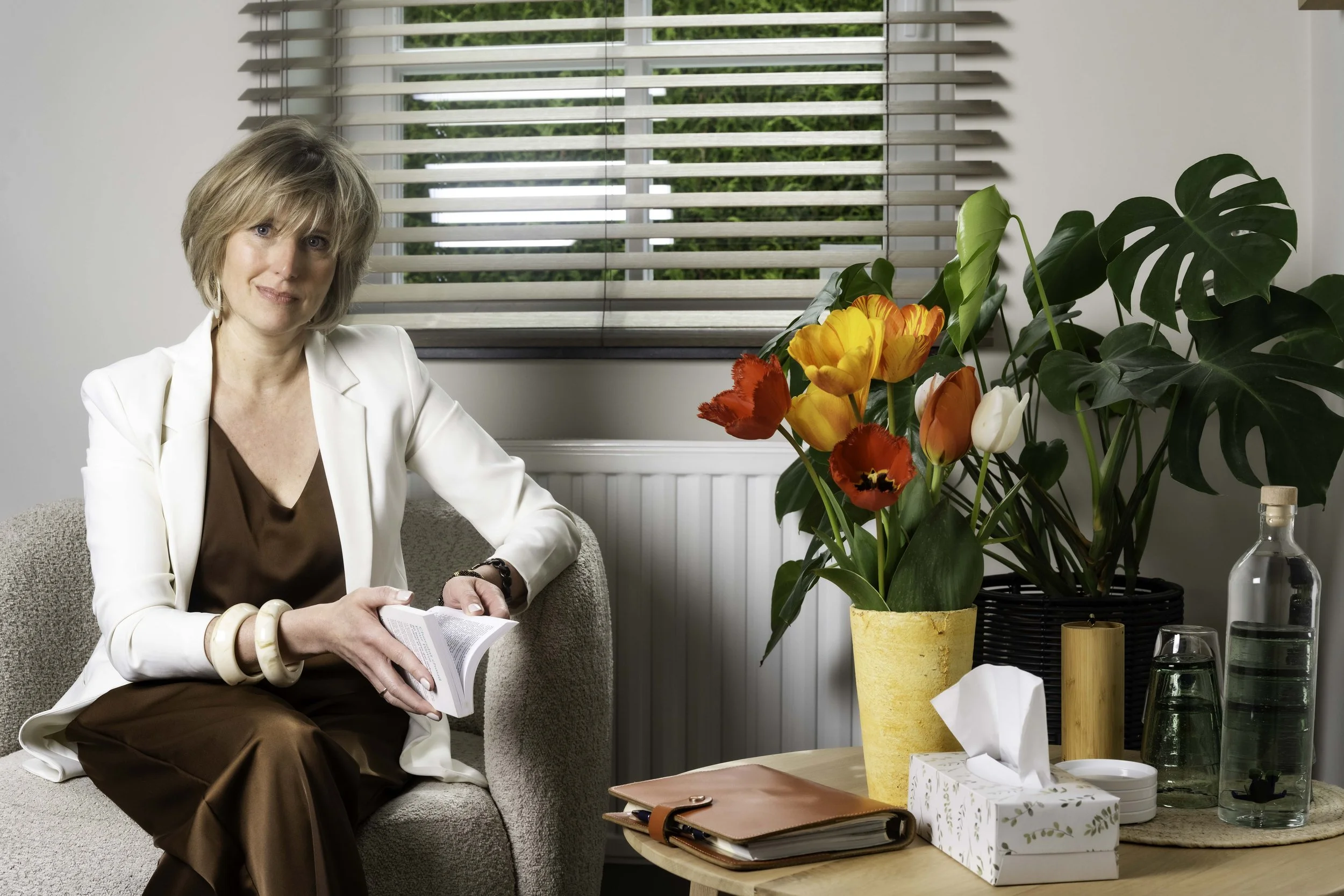 Une femme assise sur un canapé, près d'une table avec des plantes, des livres, et des objets divers, dans une pièce lumineuse avec des volets en bois.
