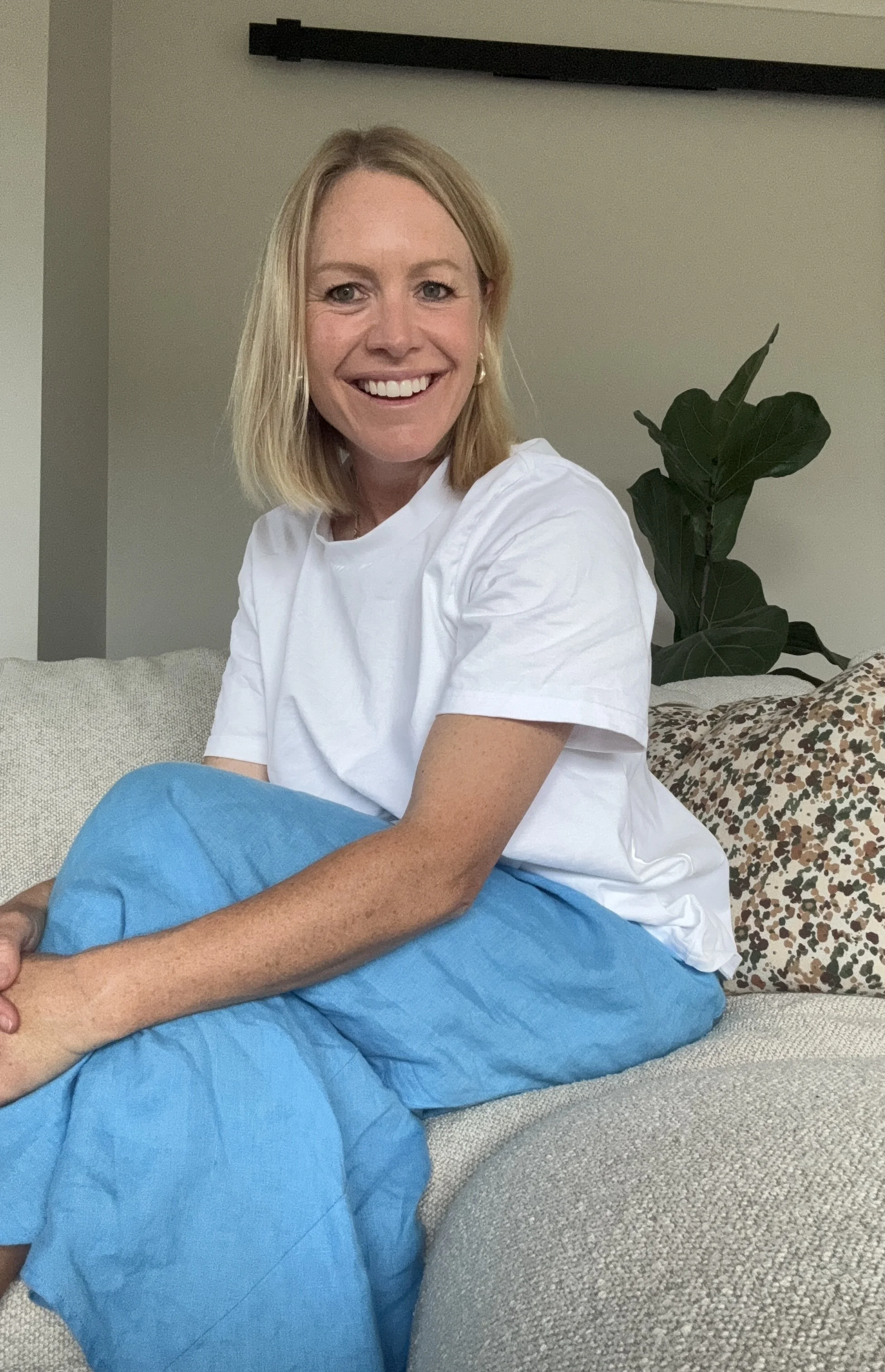 A woman with blonde hair and blue eyes smiling, sitting on a sofa with a large patterned pillow, wearing a white t-shirt and blue skirt, in a living room with a green plant behind her.