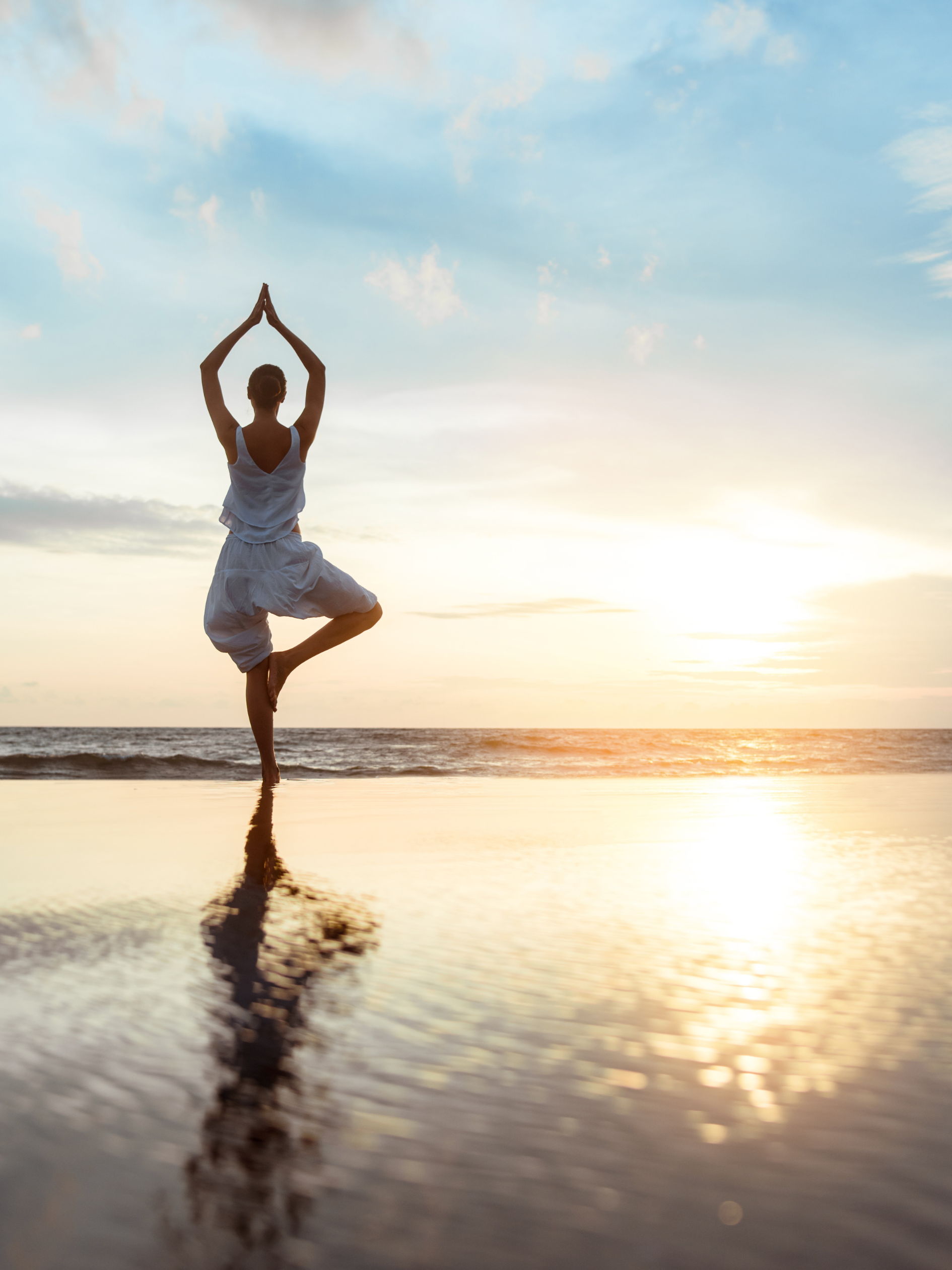 A person practicing yoga on the beach at sunset, standing on one leg in a tree pose with hands together above the head.