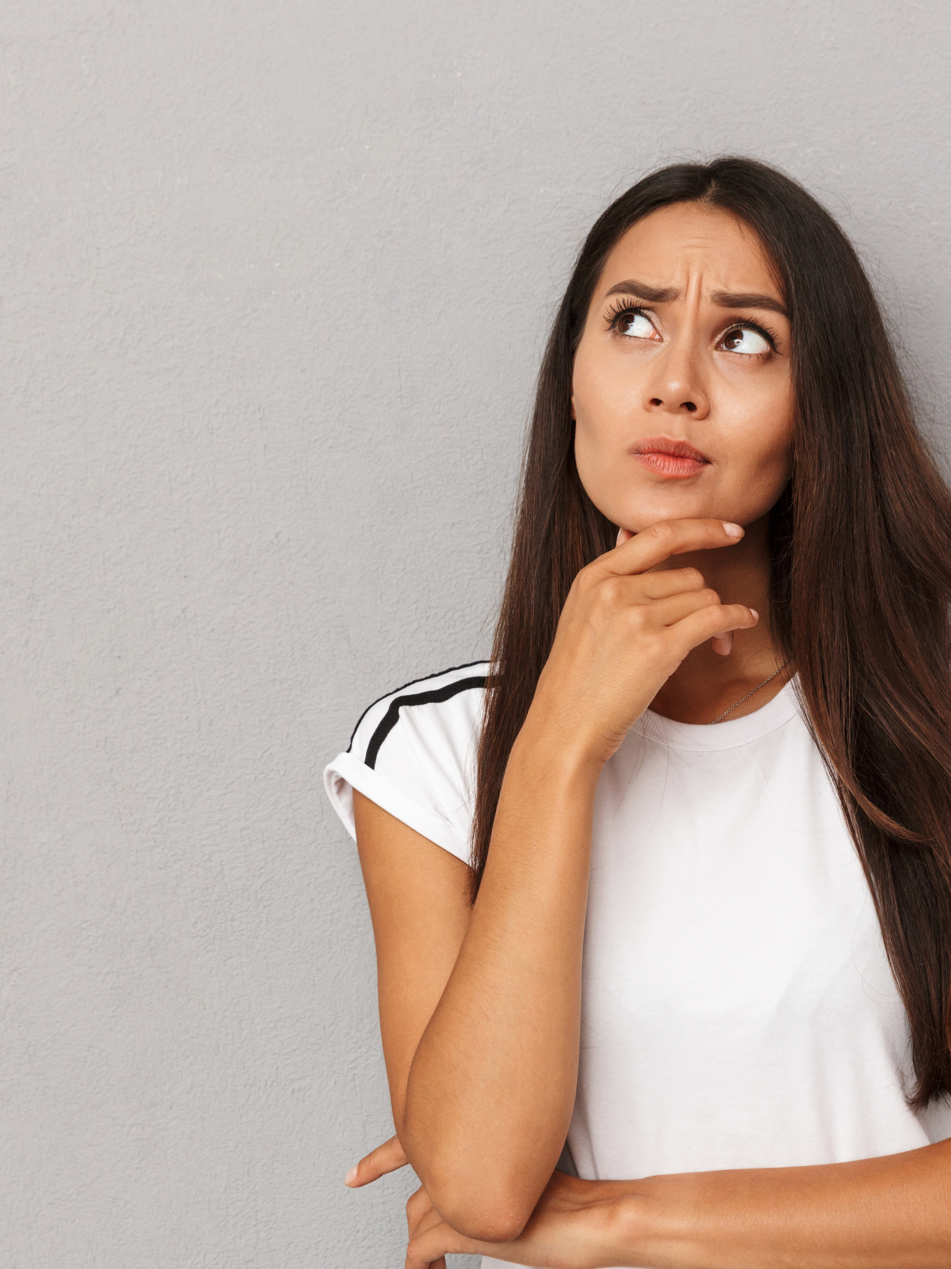 Young woman with long dark hair, wearing a white t-shirt with black stripe on shoulders, standing against a gray wall, with a puzzled or thinking expression, touching her chin with her right hand.