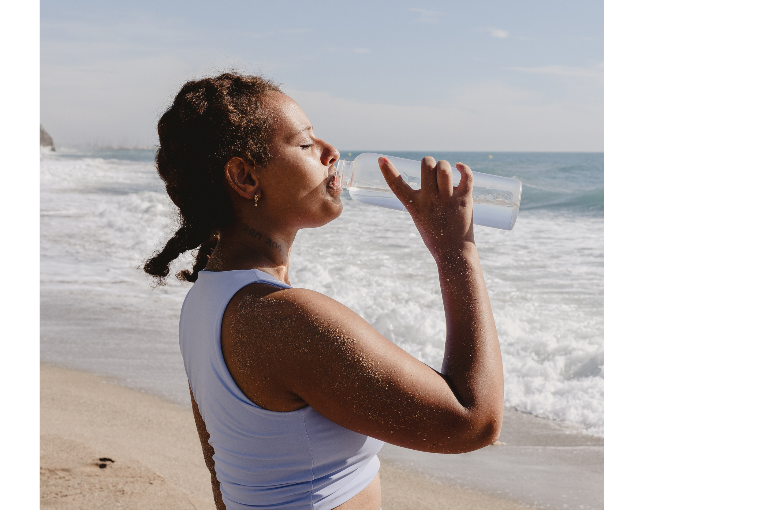 A woman on the beach drinking water from a clear water bottle with waves in the background.