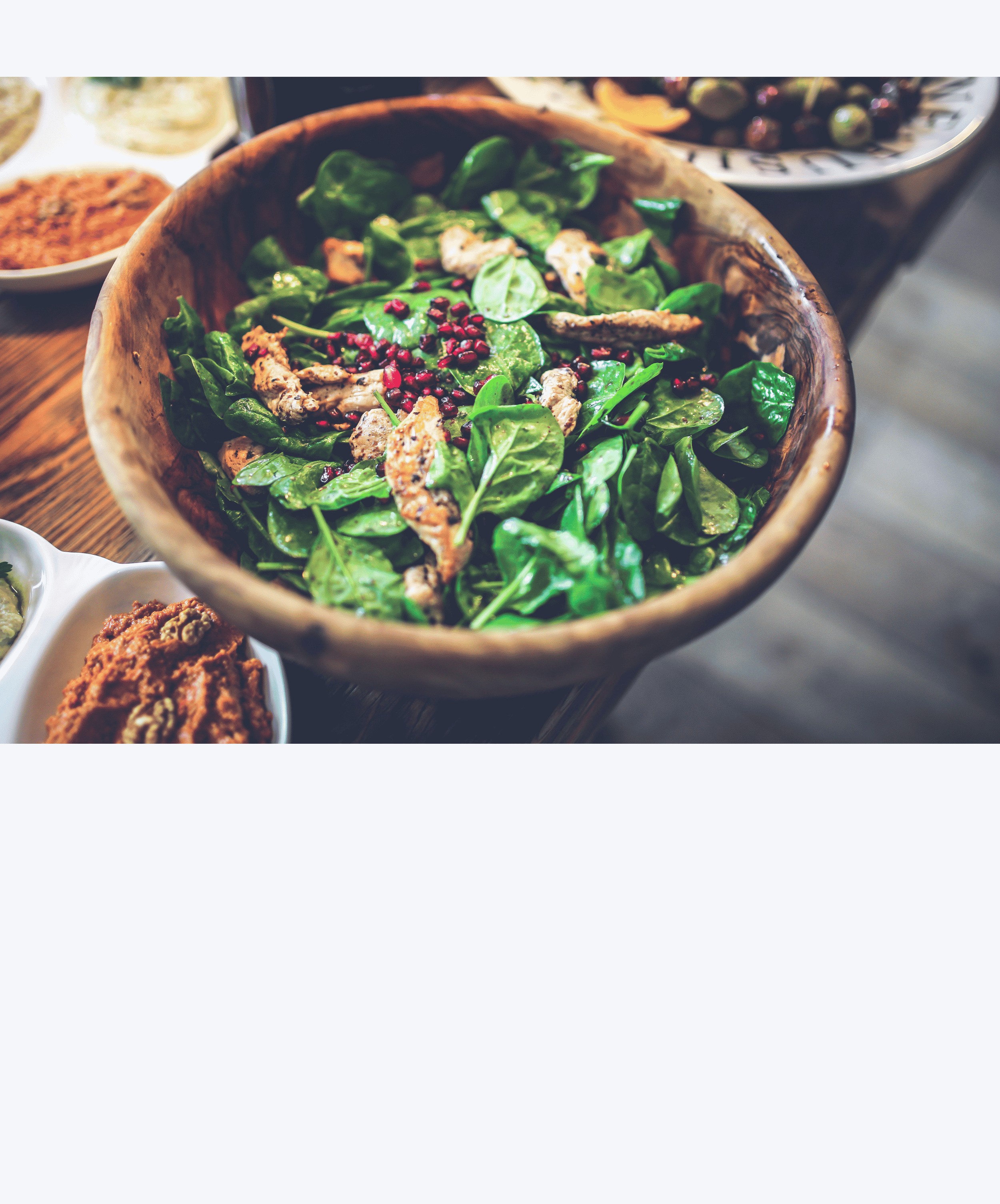 A wooden bowl filled with fresh spinach leaves, grilled chicken strips, and pomegranate seeds. In the background, there are white bowls with various ingredients or dishes on a wooden table.