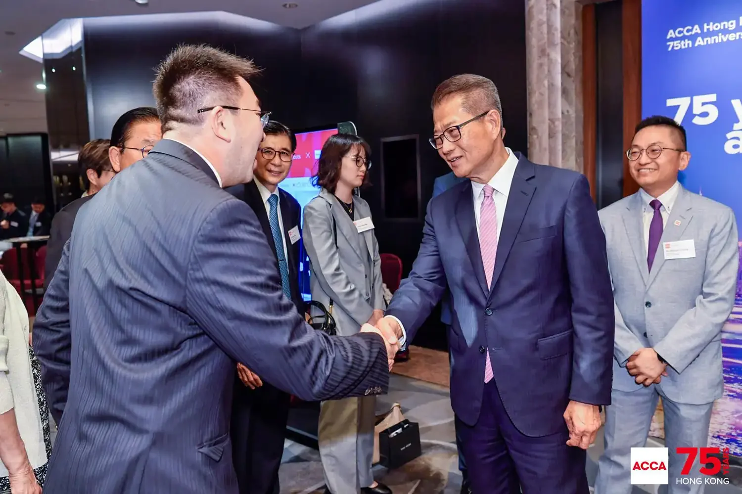 A group of professionally dressed people in suits and formal attire, attending an event, with two men shaking hands in the center. The event appears to be a celebration of the 75th anniversary of ACCA Hong Kong, as indicated by the large blue sign and logo in the background.